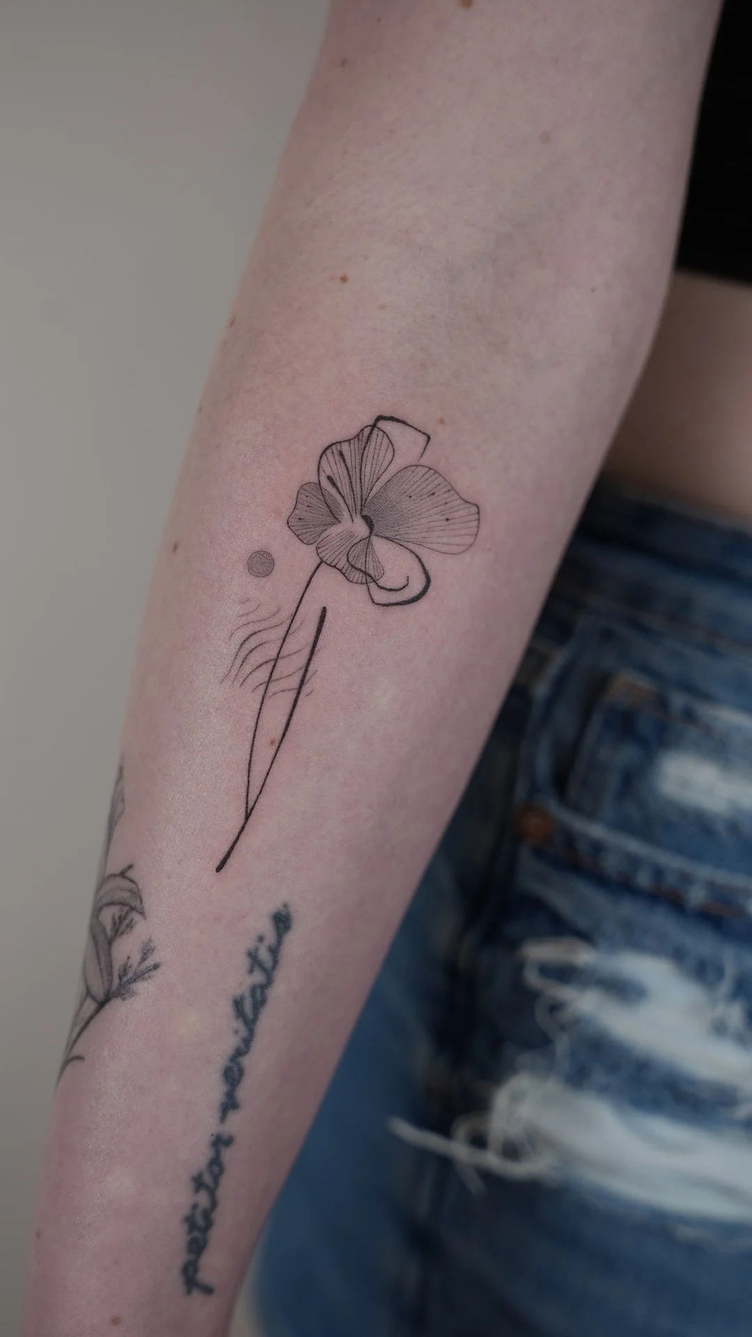 A new black ink tattoo of a minimalist flower with linear petals on a person's forearm, with a small dot and curved lines nearby, and part of a script tattoo below.