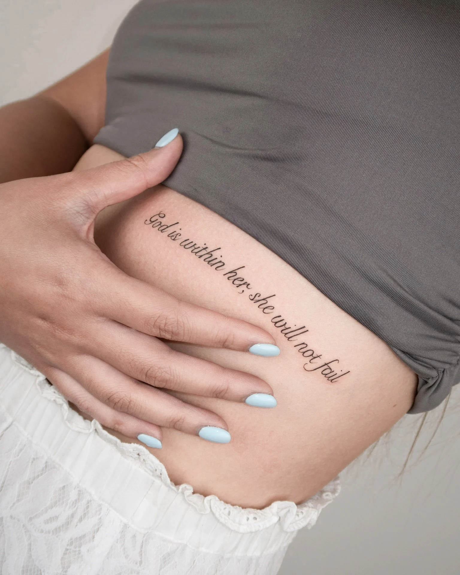 A person's side torso with a tattoo that reads 'God is within her, she will not fail' in cursive script, partially covered by a gray top and white lace skirt, with hand and nails visible.