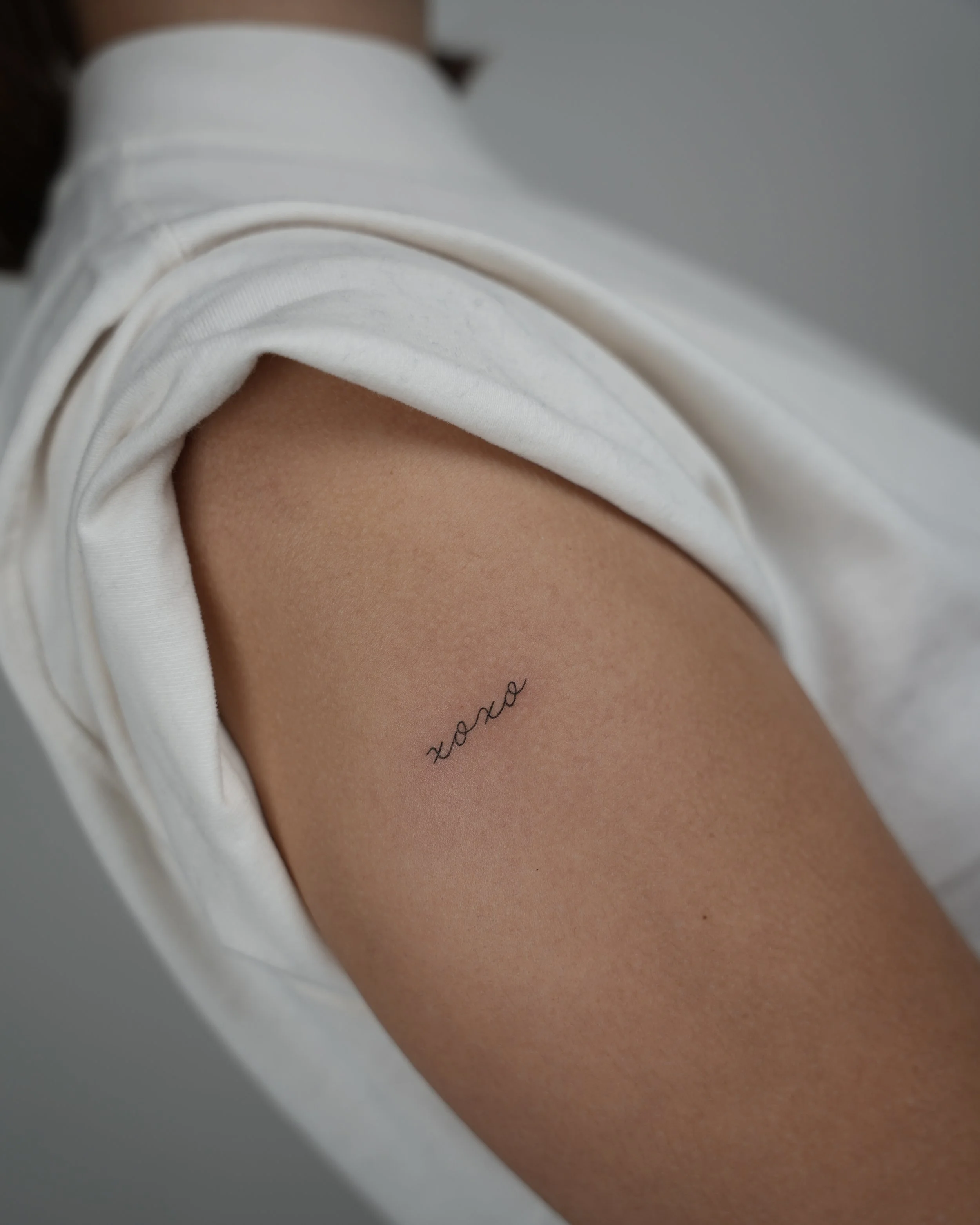 A close-up of a person's upper arm with a small, elegant black tattoo that reads 'xoxo'. The person is wearing white clothing, and the background appears to be a neutral wall.