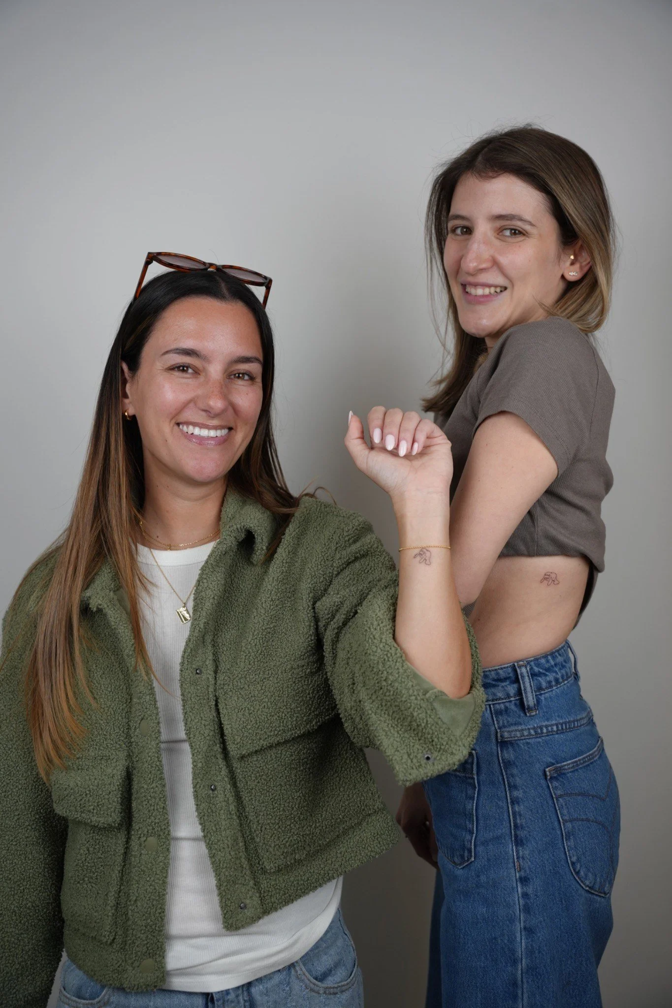 Two women smiling and posing together against a plain gray background with new fineline tattoos created at UPLIFT tattoo and piercing
