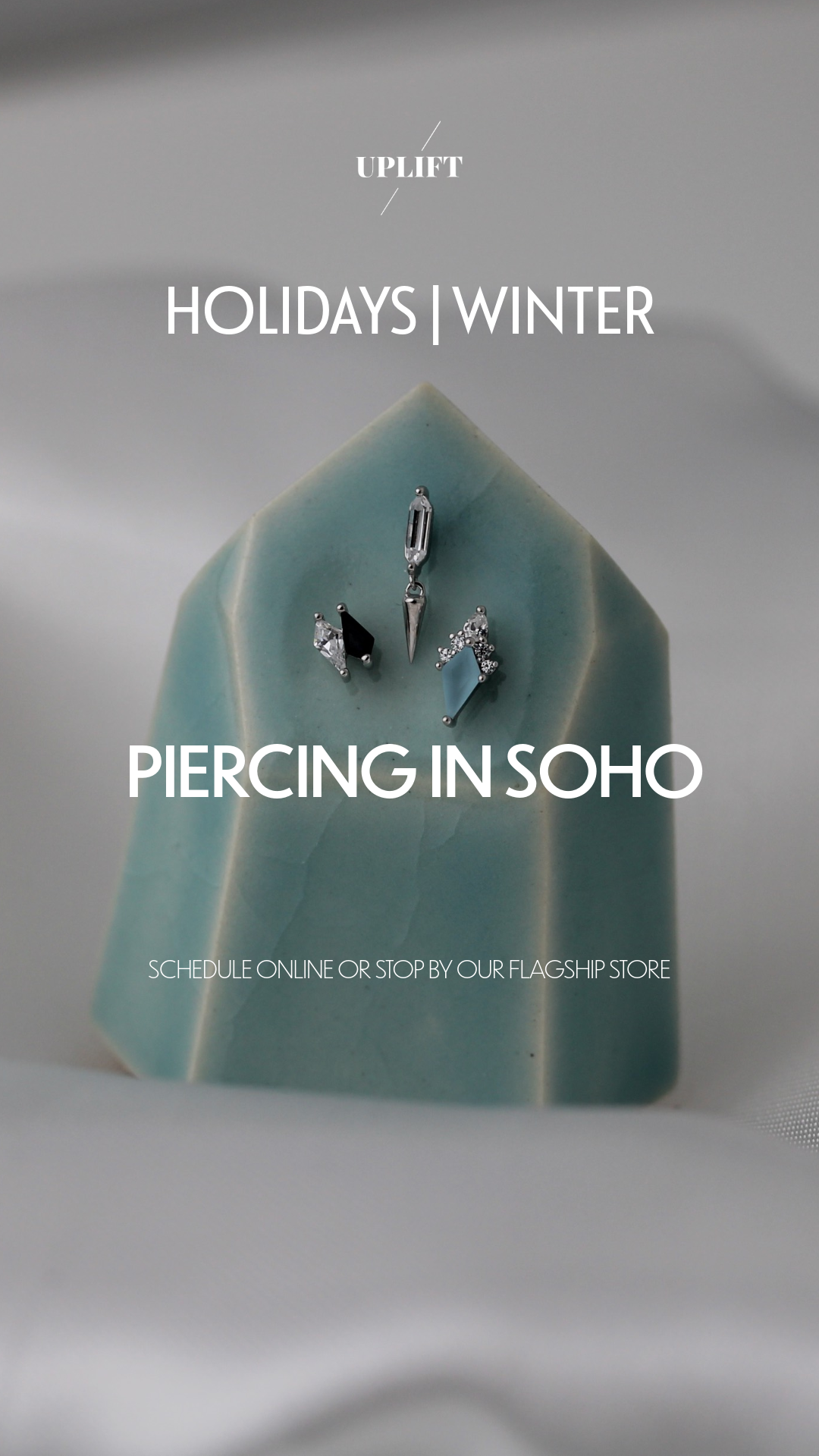 Jewelry display with three earrings on a light blue geometric stone, promoting holiday winter piercing in Soho, with a call to schedule online or visit the flagship store.