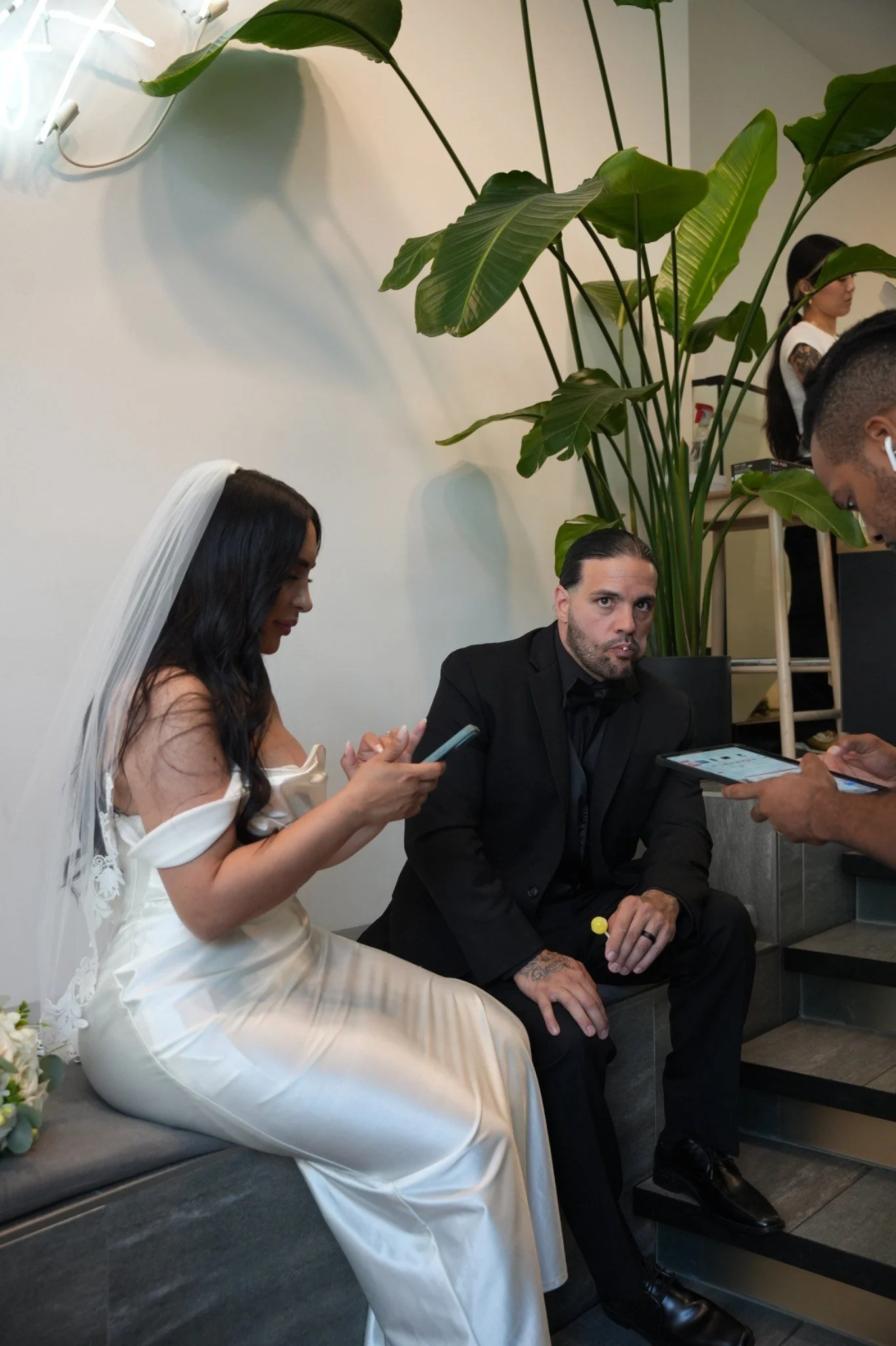 A bride and groom sitting on a bench, with the bride wearing a white wedding dress and veil, and the groom in a black suit, surrounded by onlookers using their phones.