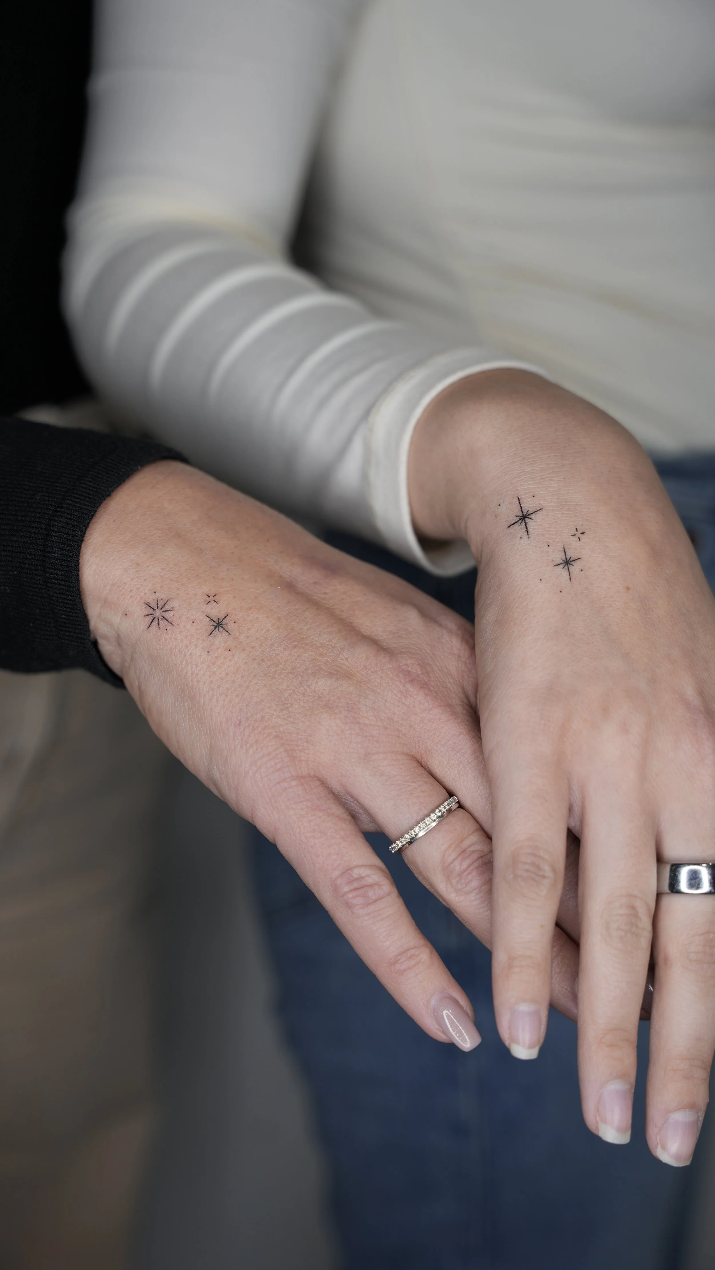 Close-up of two hands with matching star-shaped tattoos, one wearing a black long-sleeve shirt and the other a white long-sleeve shirt, with rings on their fingers.