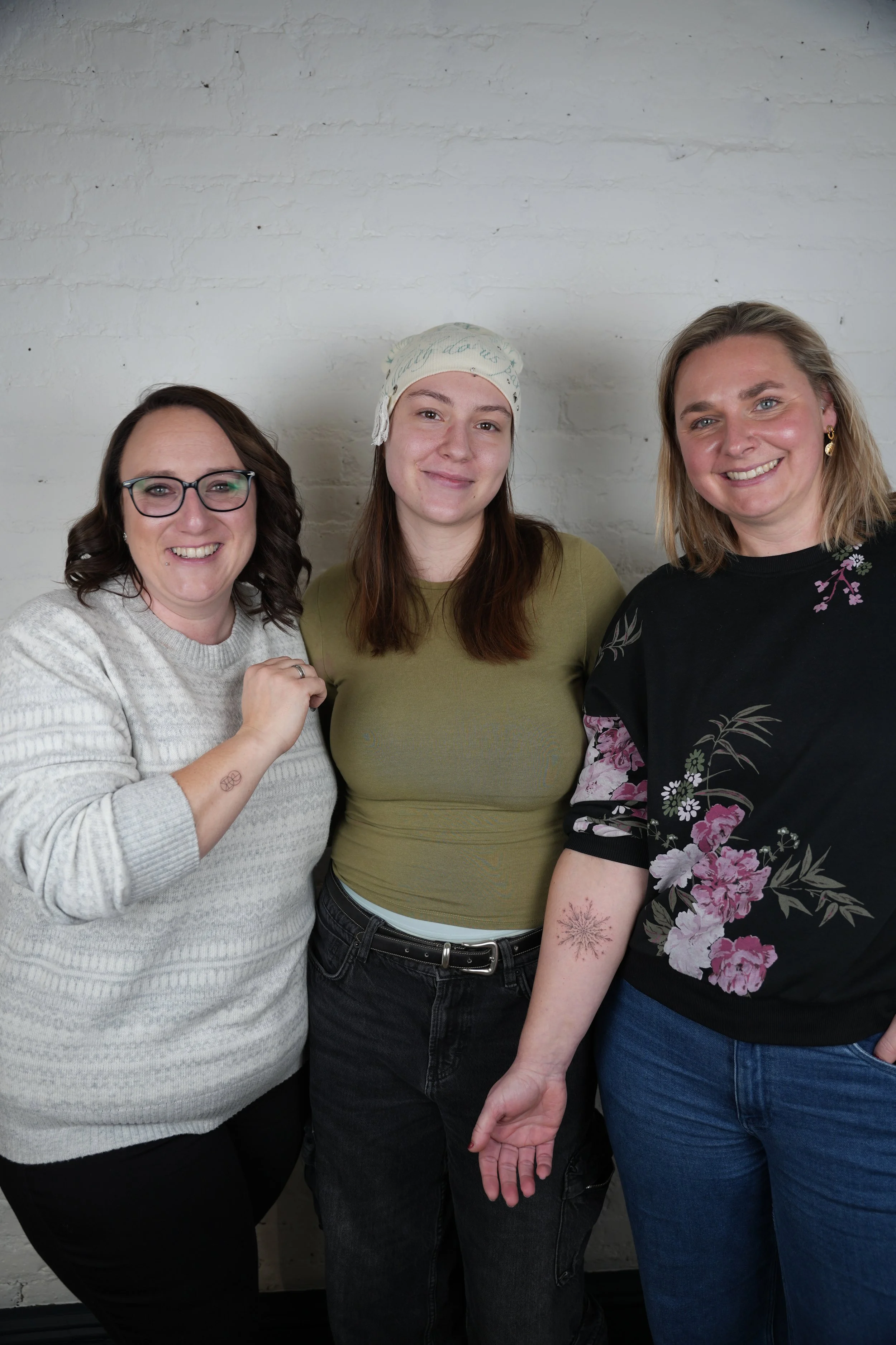 Three smiling women standing together against a white brick wall. The woman on the left has short dark hair, glasses, and is wearing a light gray sweater. The woman in the middle has long brown hair, a light-colored headscarf, and a green top. The wo