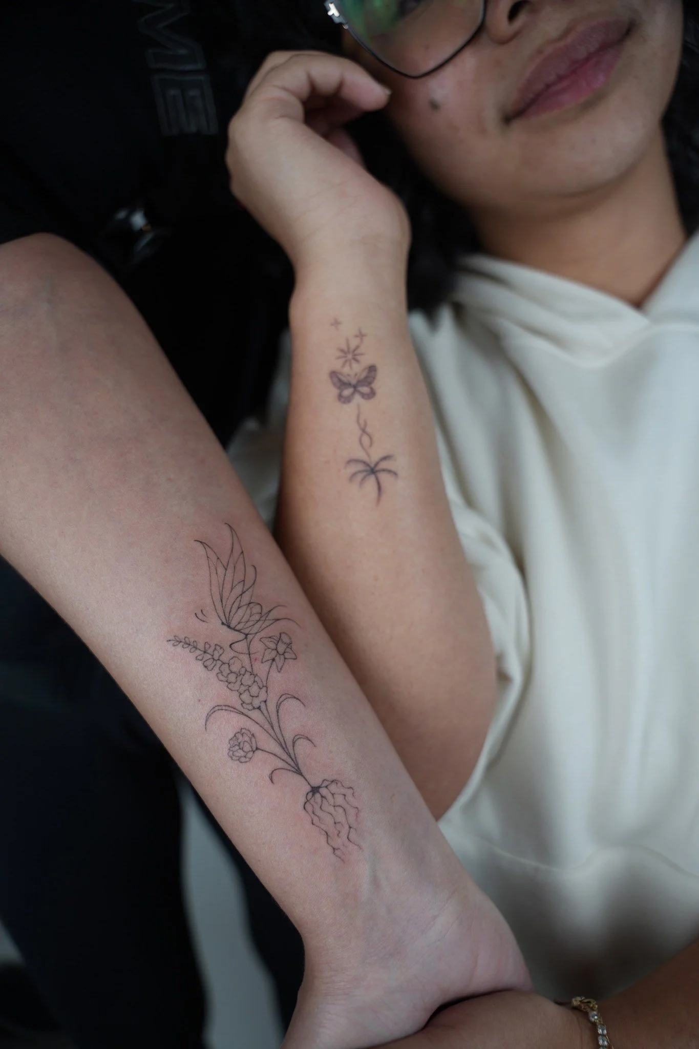 Close-up of a woman's arm with a floral and butterfly tattoo, resting on another person's arm. The woman is wearing glasses, a white hoodie, and a gold bracelet. Their faces are partially visible.