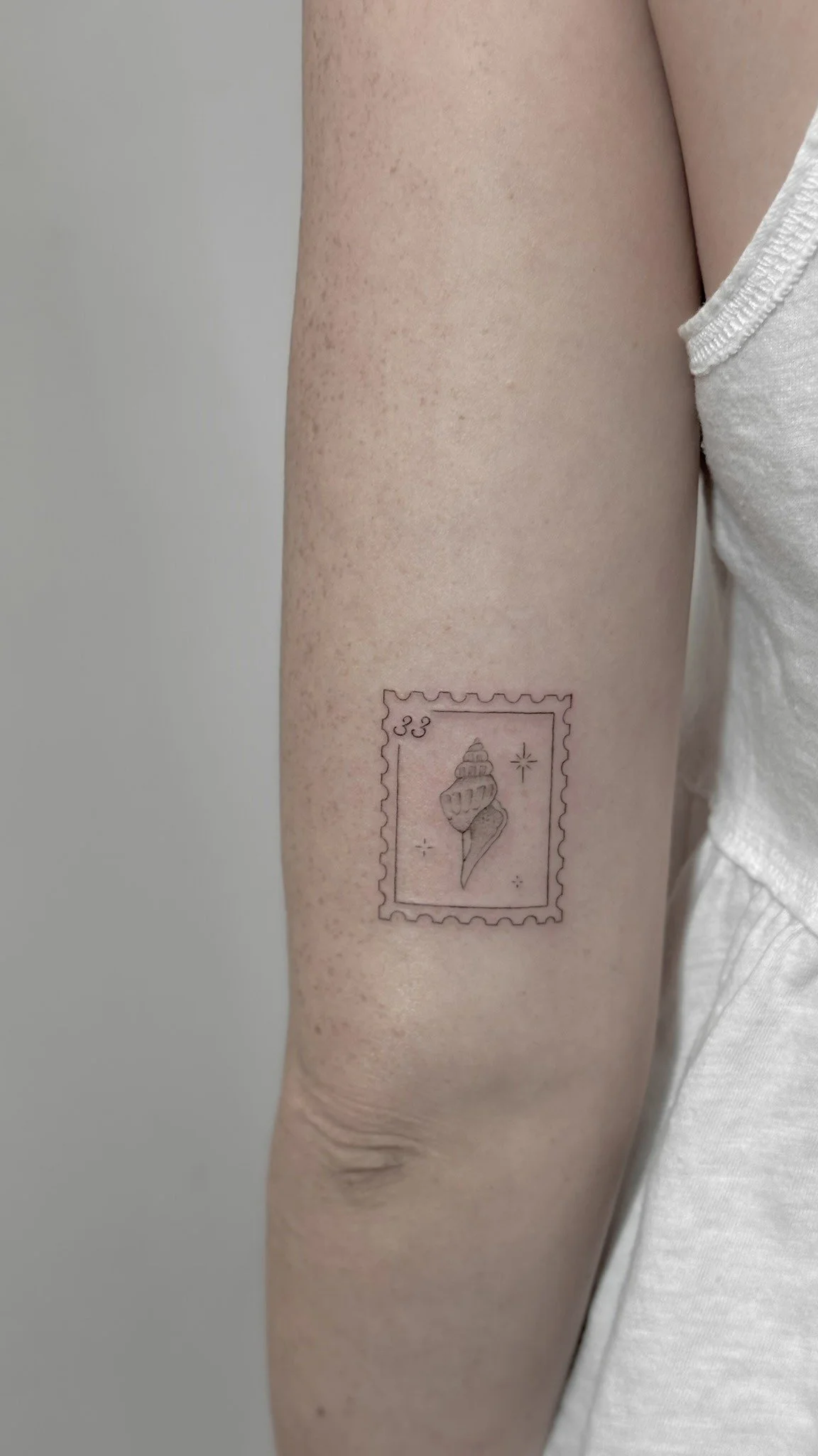 A person's forearm with a tattoo of a postage stamp featuring a shell and stars.