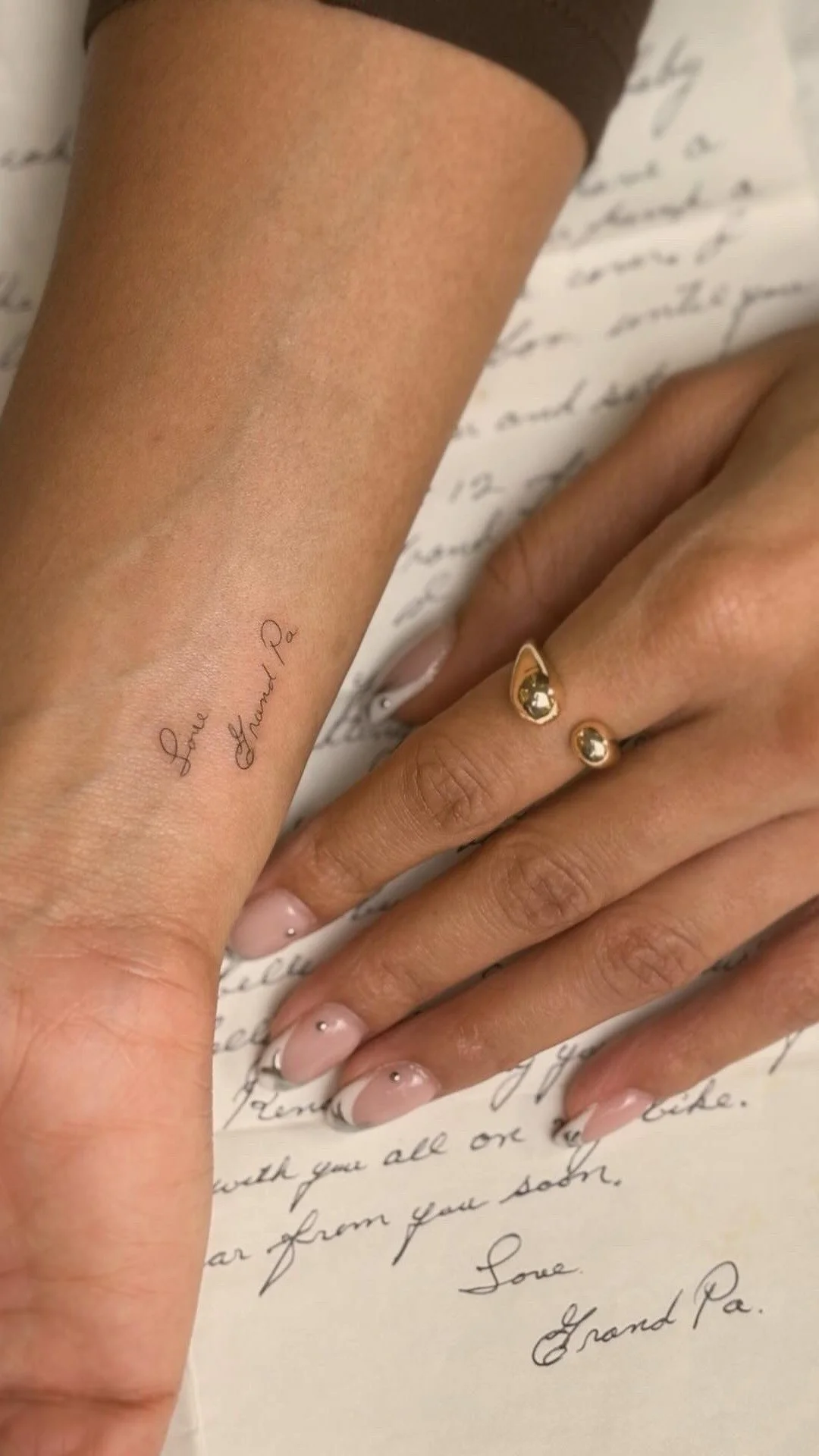Close-up of two hands, one with a small tattoo reading 'Love Grandma Pa' on the wrist and the other with a gold ring and manicured nails, resting on handwritten letter paper.