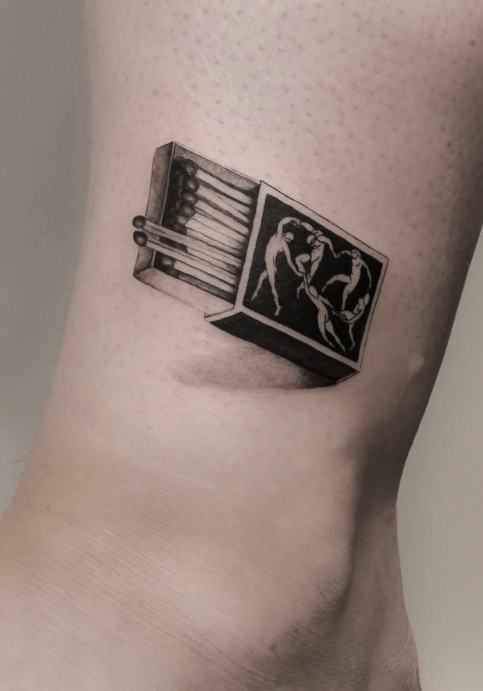 Black and gray tattoo of a box of matches and a box with dancing figures on skin.
