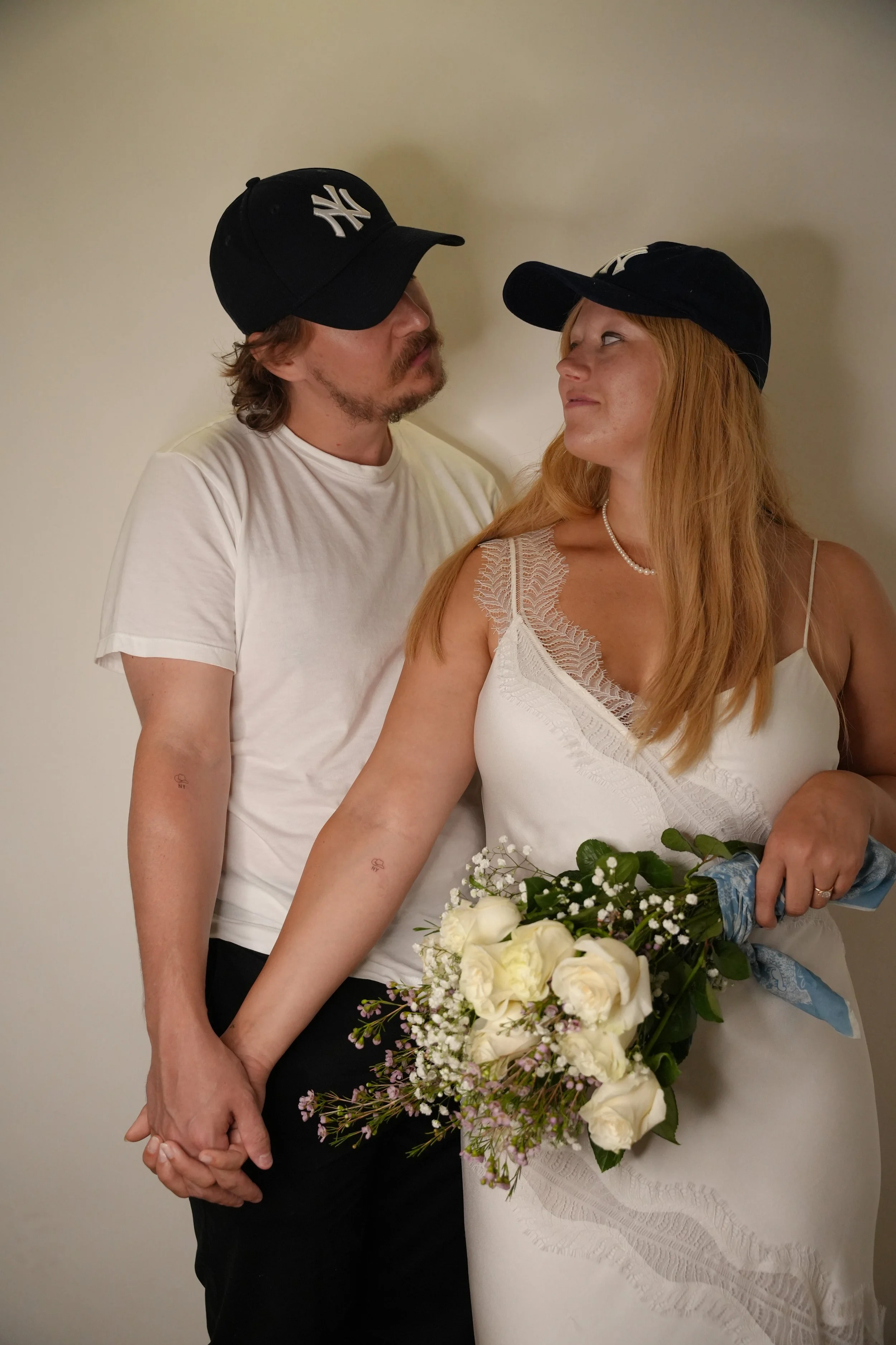 A couple holding hands, with the woman holding a bouquet of white roses and greenery, both wearing baseball caps, standing against a plain wall.
