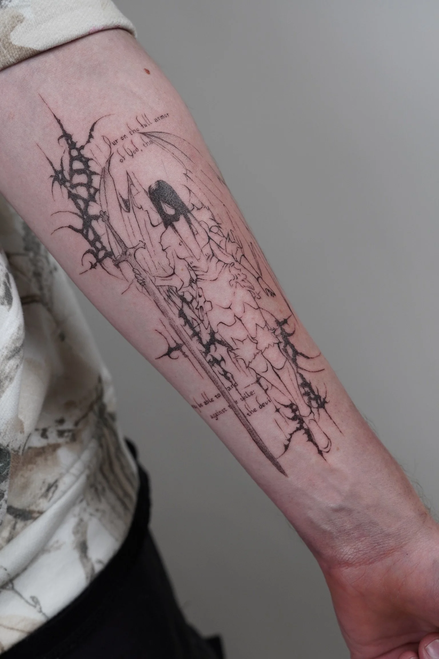 Line art tattoo on person's forearm depicting a woman with a sword and sword's shadow, with some text around the image.