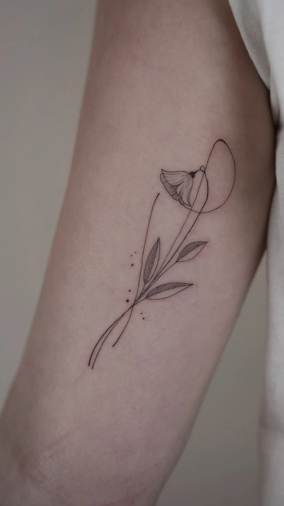 Minimalist black line tattoo of a flower with leaves and a butterfly on a person's upper arm.