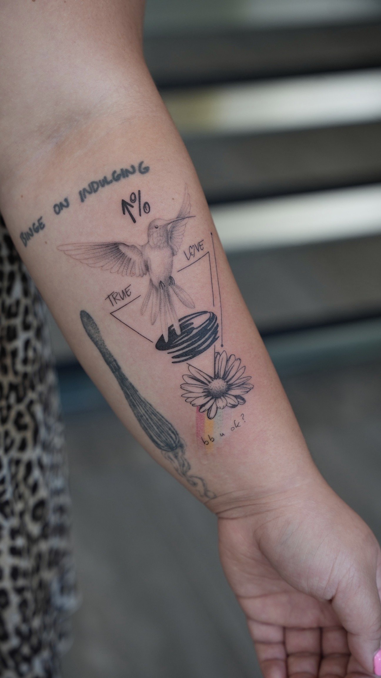 Tattoo of a hummingbird with the phrase 'since on indulging 1%' and a flower on a person's arm.