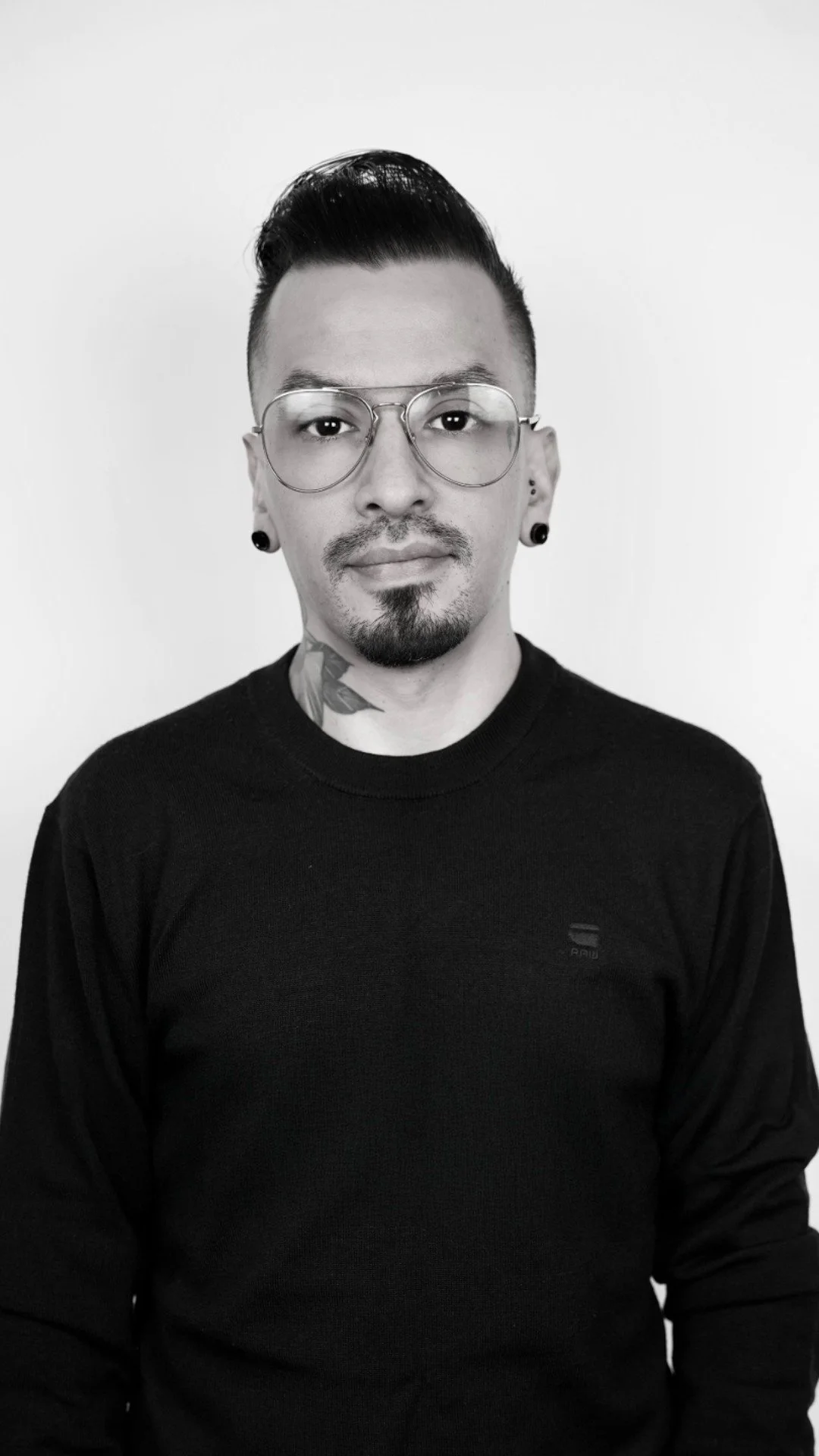 Black and white portrait of a man with glasses, a short beard, dyed hair, earrings, and a tattoo on his neck, wearing a black sweater.