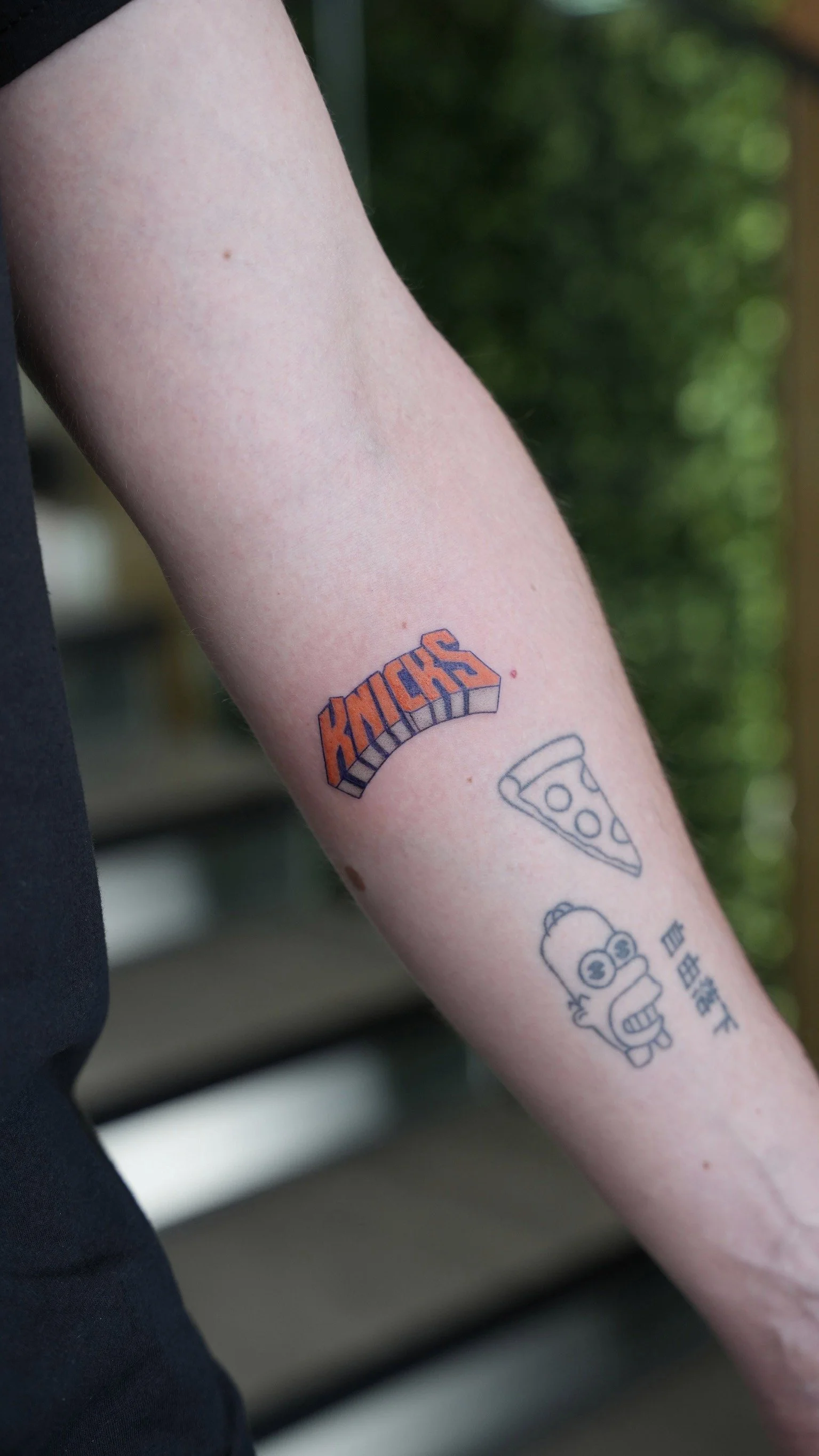 Tattoo of the New York Knicks logo on a person's forearm, with additional tattoos of a slice of pizza, a cartoon character, and some Chinese characters.