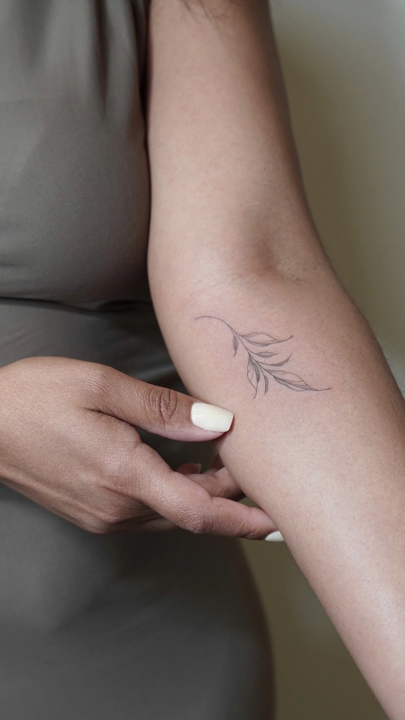 Close-up of a person's arm with a delicate black ink tattoo of a branch with leaves, and a hand with light-colored nail polish gently touching the arm.