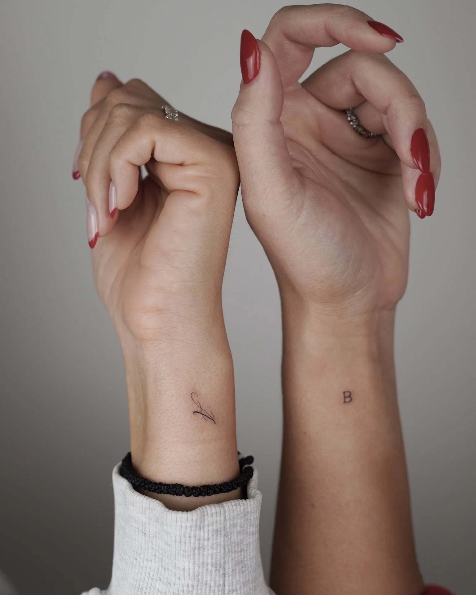 Two hands with red manicured nails and tattoos on the wrists, posed together against a neutral background.