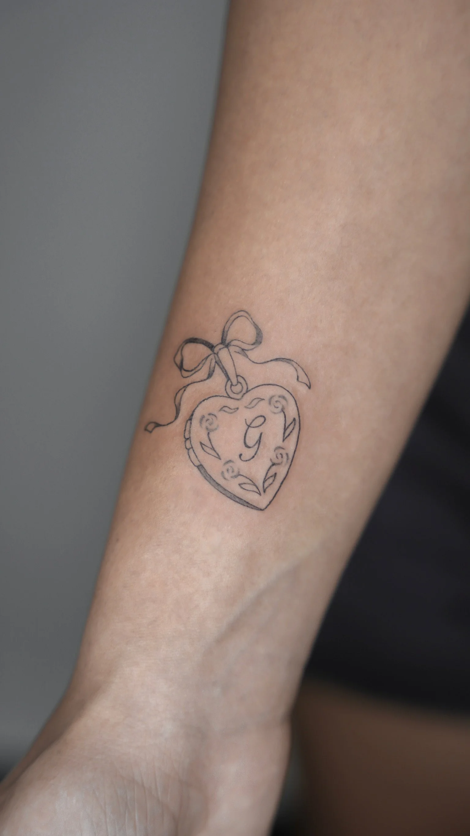 Close-up of a tattoo of a heart-shaped jewelry tag with a bow on top, featuring an ornate letter G in the center on a person's arm.