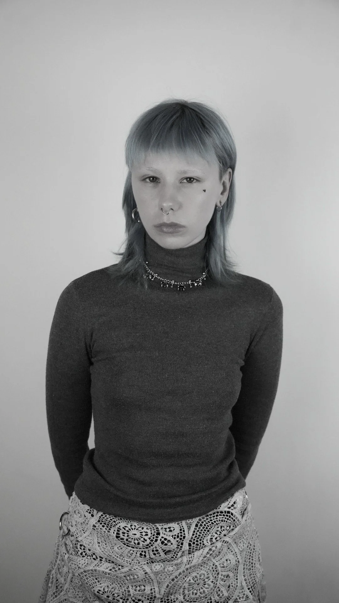 A woman with short, multi-colored hair, wearing a dark turtleneck top and patterned skirt, adorned with earrings, a necklace, a septum piercing, and a small heart tattoo on her cheek, standing against a plain background.