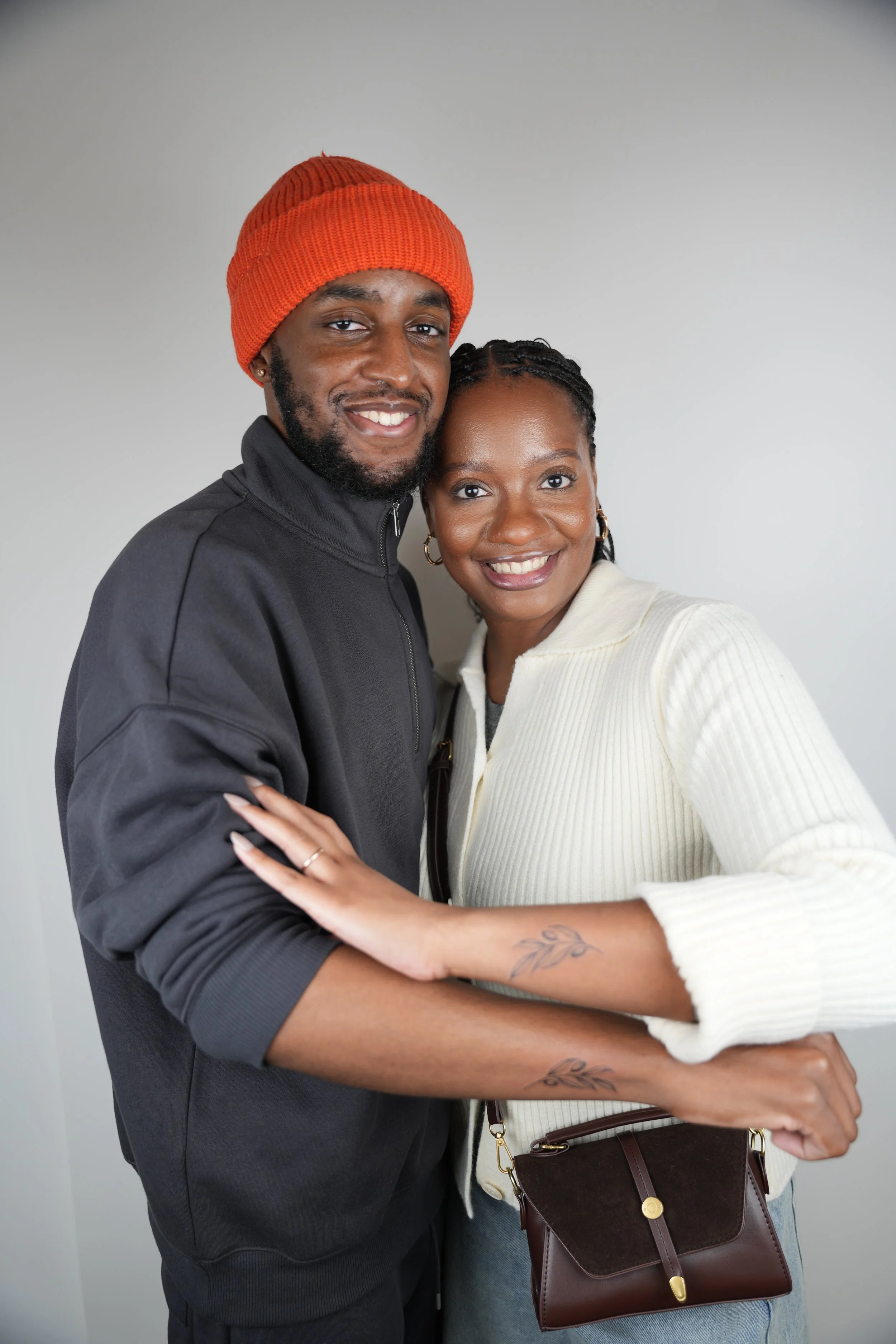 A smiling Black man wearing an orange beanie and black jacket hugging a smiling Black woman with braided hair, wearing a cream sweater and carrying a brown purse, against a plain light gray background.