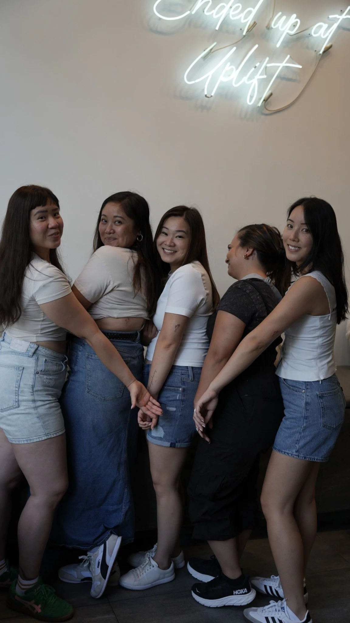A group of five women standing together, holding hands, in front of a white wall with a neon sign that says "Ended up at Upgrade."