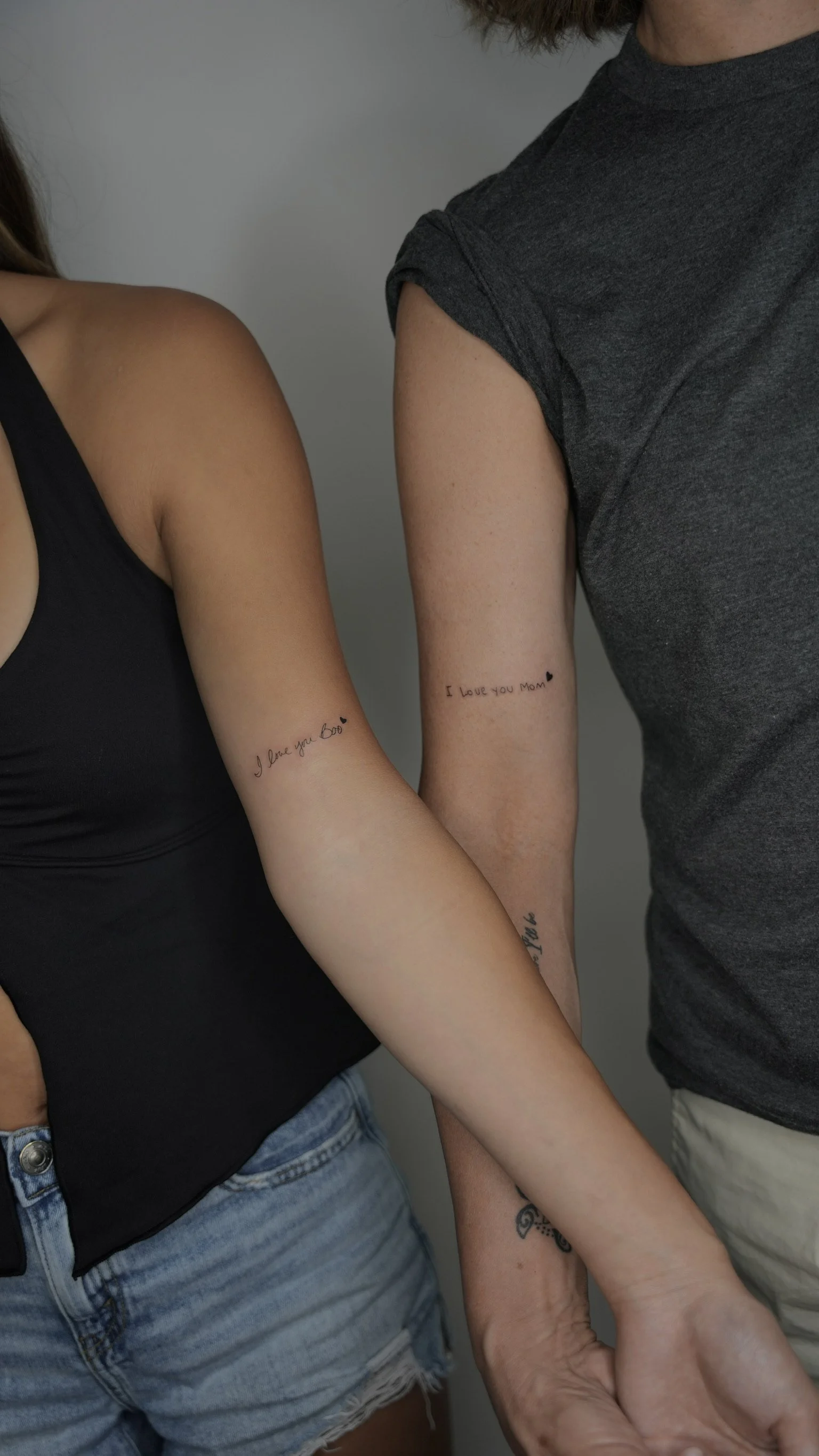 Two women with matching tattoos on their arms that say, "I love you Boo" and "I love you Mom" with small heart icons.