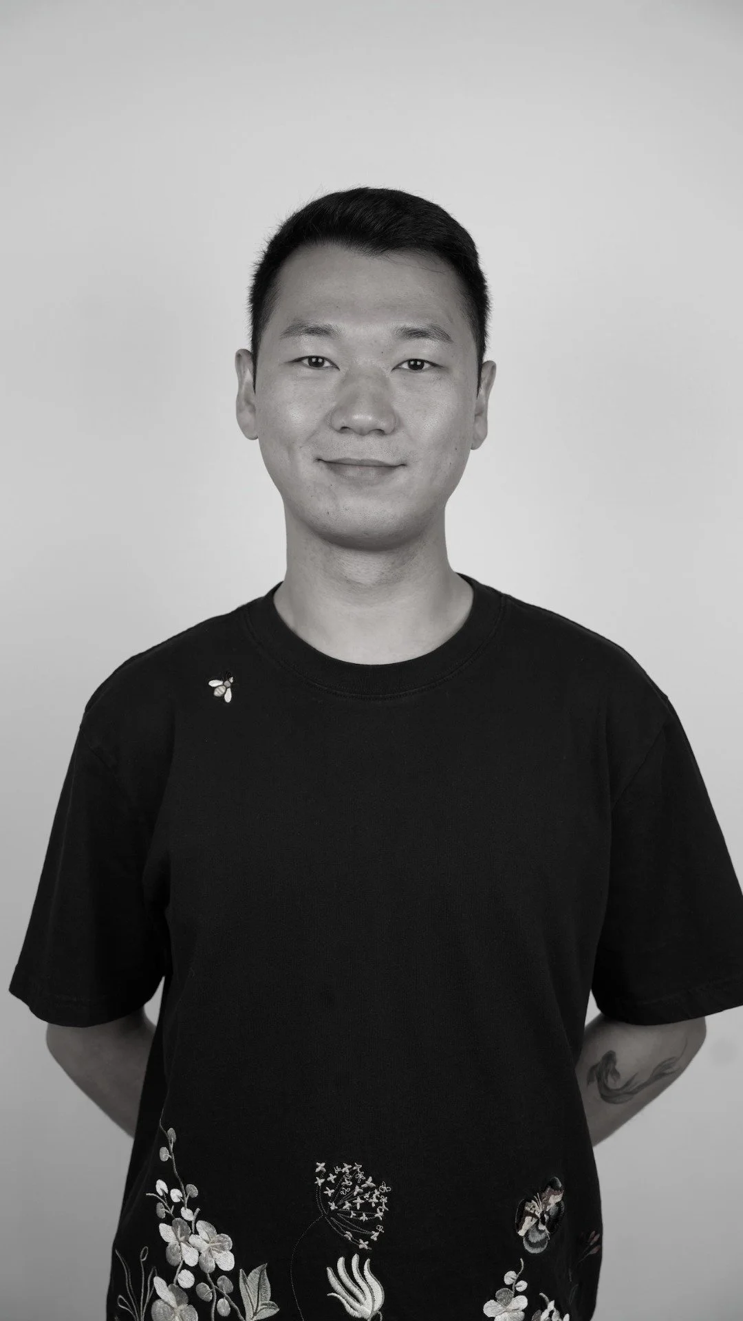 Black and white portrait of a young Asian man with short hair, wearing a black t-shirt with floral designs and a small bee graphic on the shoulder, standing against a plain background.