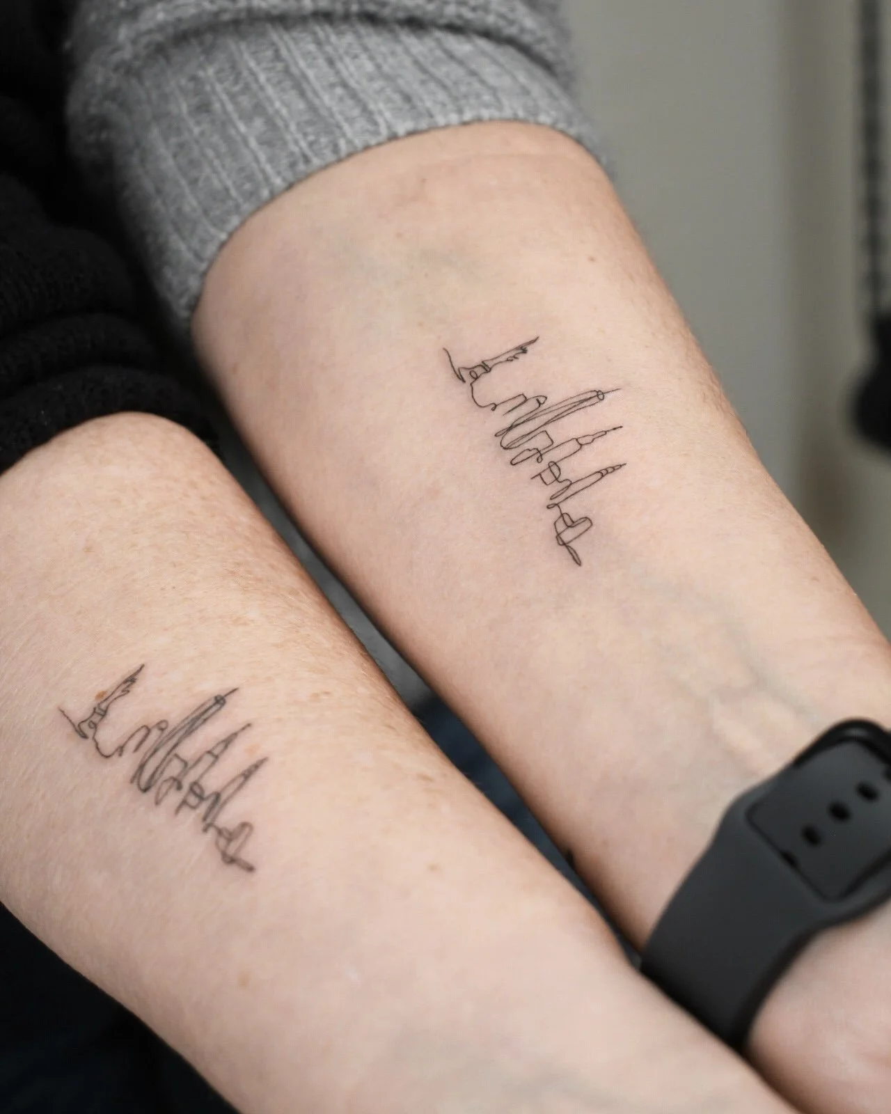 Two forearms with matching tattoos of the word 'Family' written in a continuous line in cursive style.