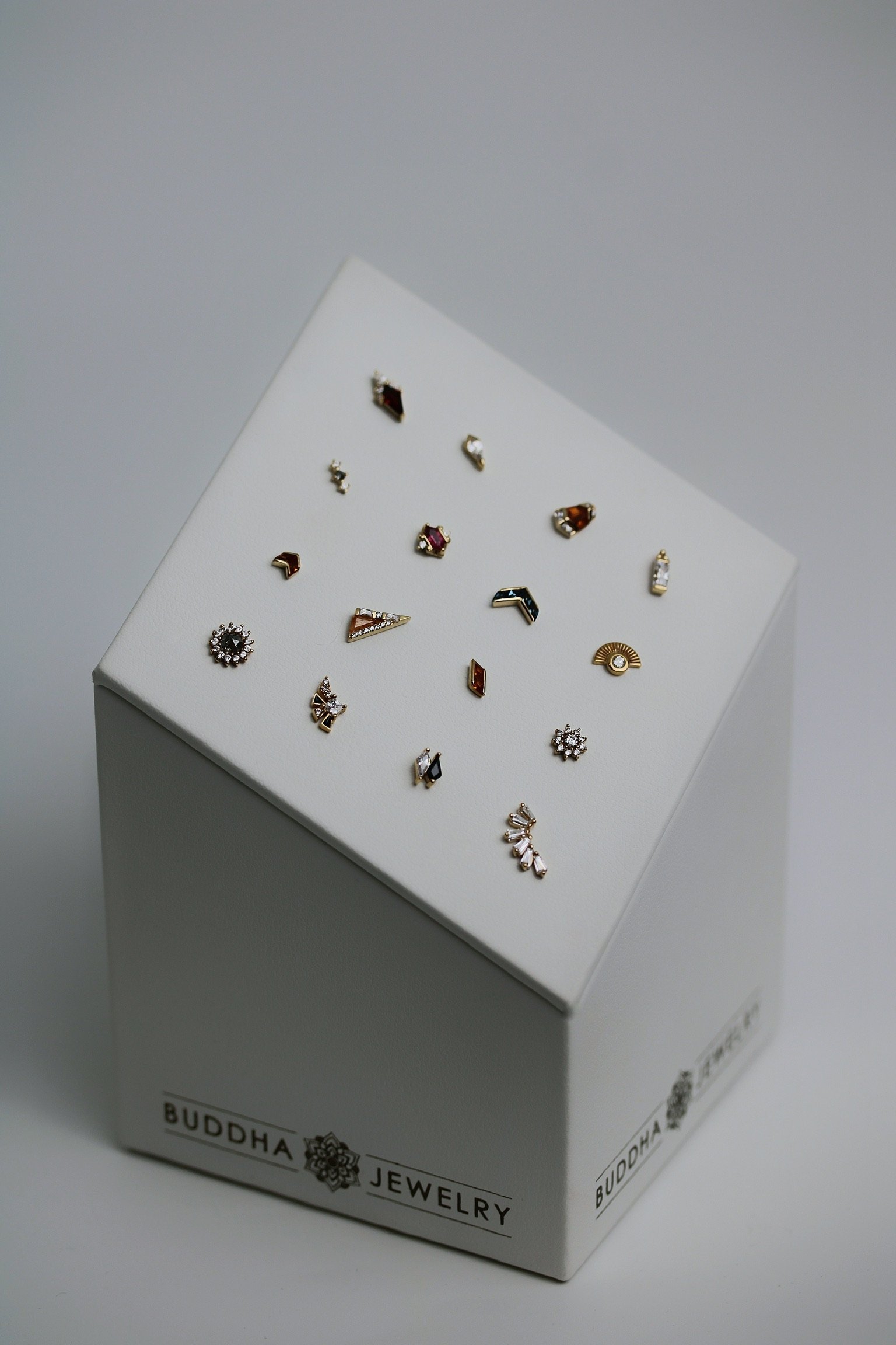 Close-up view of a white box titled 'Buddha Jewelry' containing an assortment of small, colorful, decorative jewelry pieces with gemstones and intricate designs.