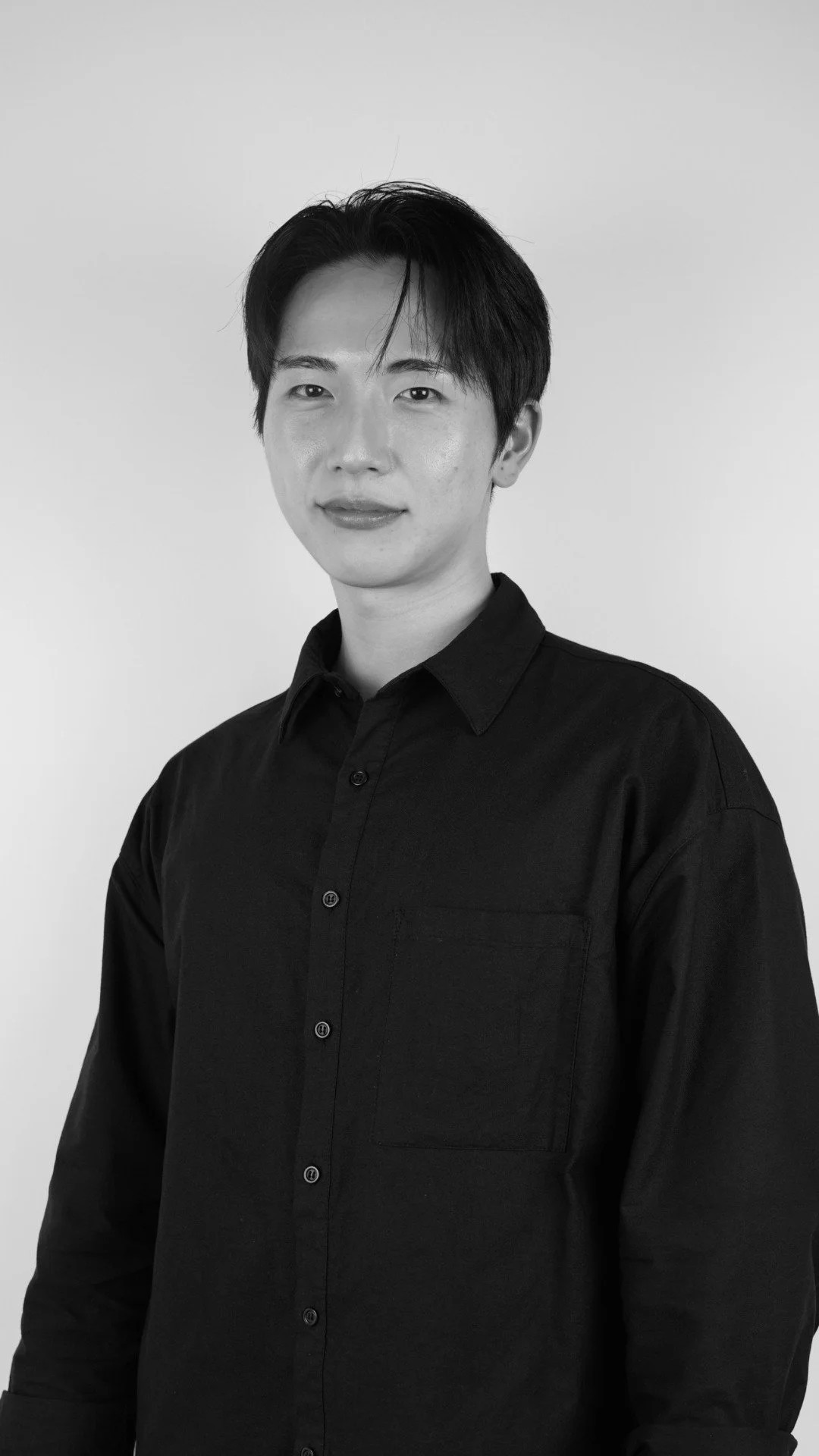 Black and white portrait of a woman with short black hair, wearing a dark button-up shirt, standing against a plain background.