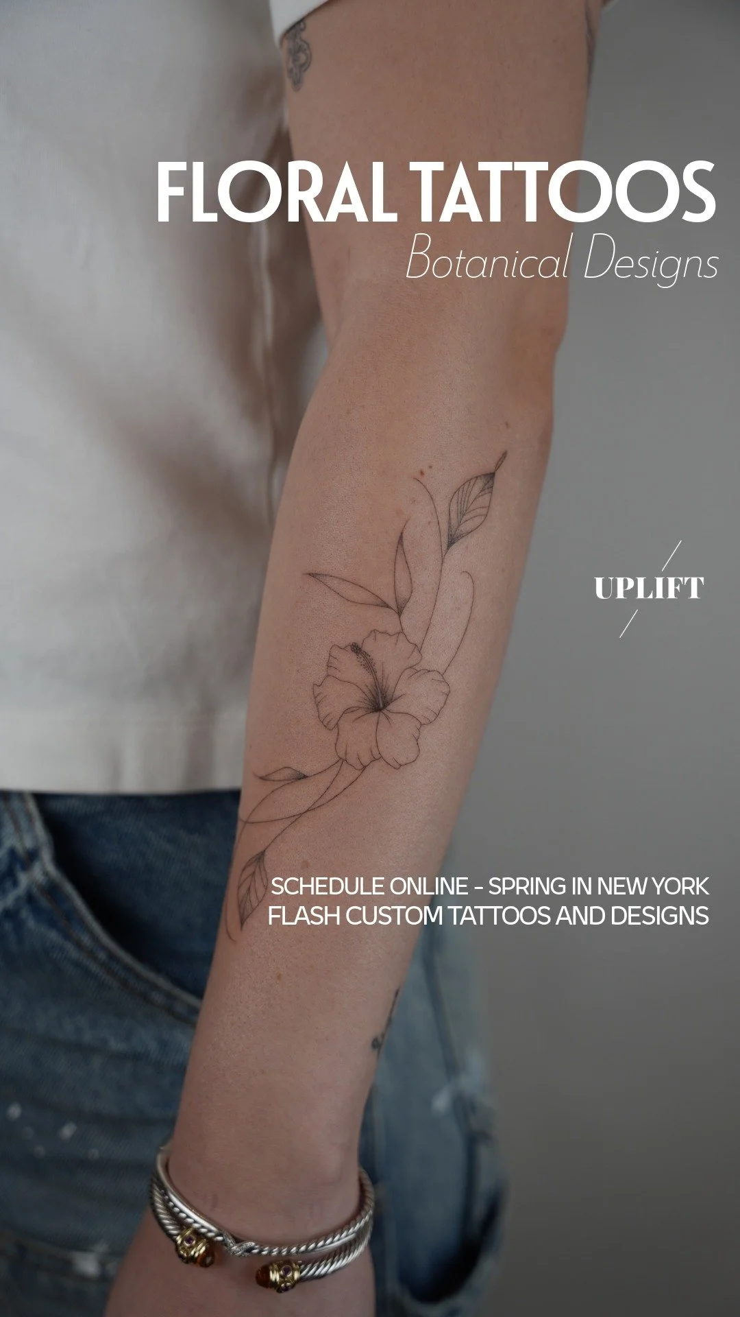 A person's forearm with a floral tattoo of a hibiscus flower and leaves in delicate, black ink outlines. The person is wearing a silver bracelet on the wrist. The background is neutral and blurred.