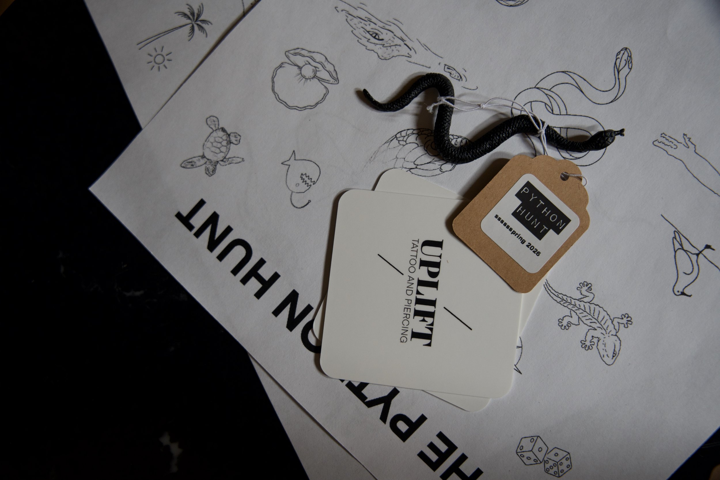 A black and brown crawling snake placed on a sheet of paper with black line drawings of animals, including a turtle and gecko, and text that says 'NEON HUNT'. There are two tags attached to the snake; one says 'PYTHON HUNT' and the other says 'UPLIFT TATTOO AND PIERCING'.
