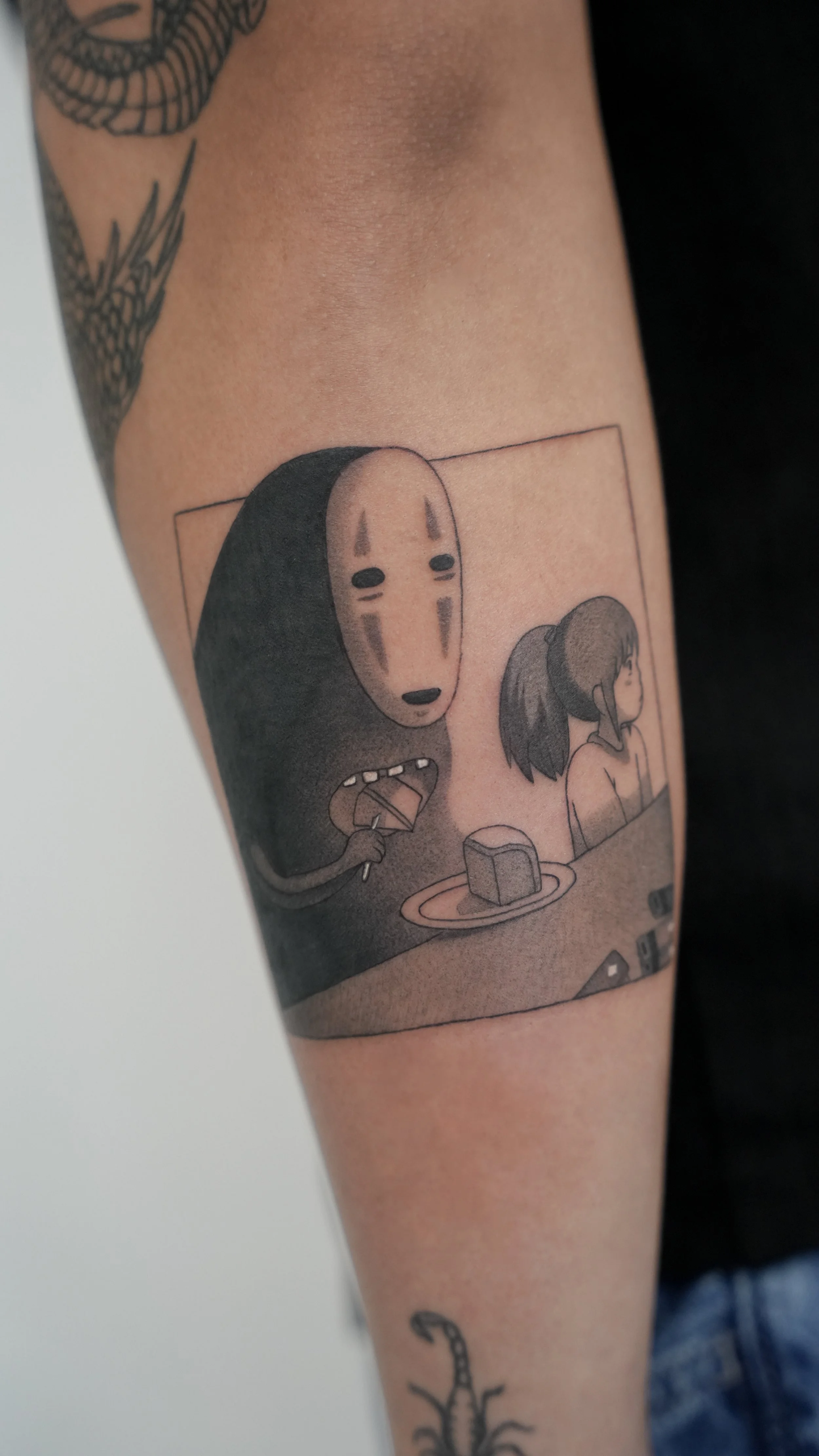 Tattoo of a scene from the animated film 'Spirited Away' featuring No-Face and a young girl sitting at a table with a square-shaped pastry on a plate.