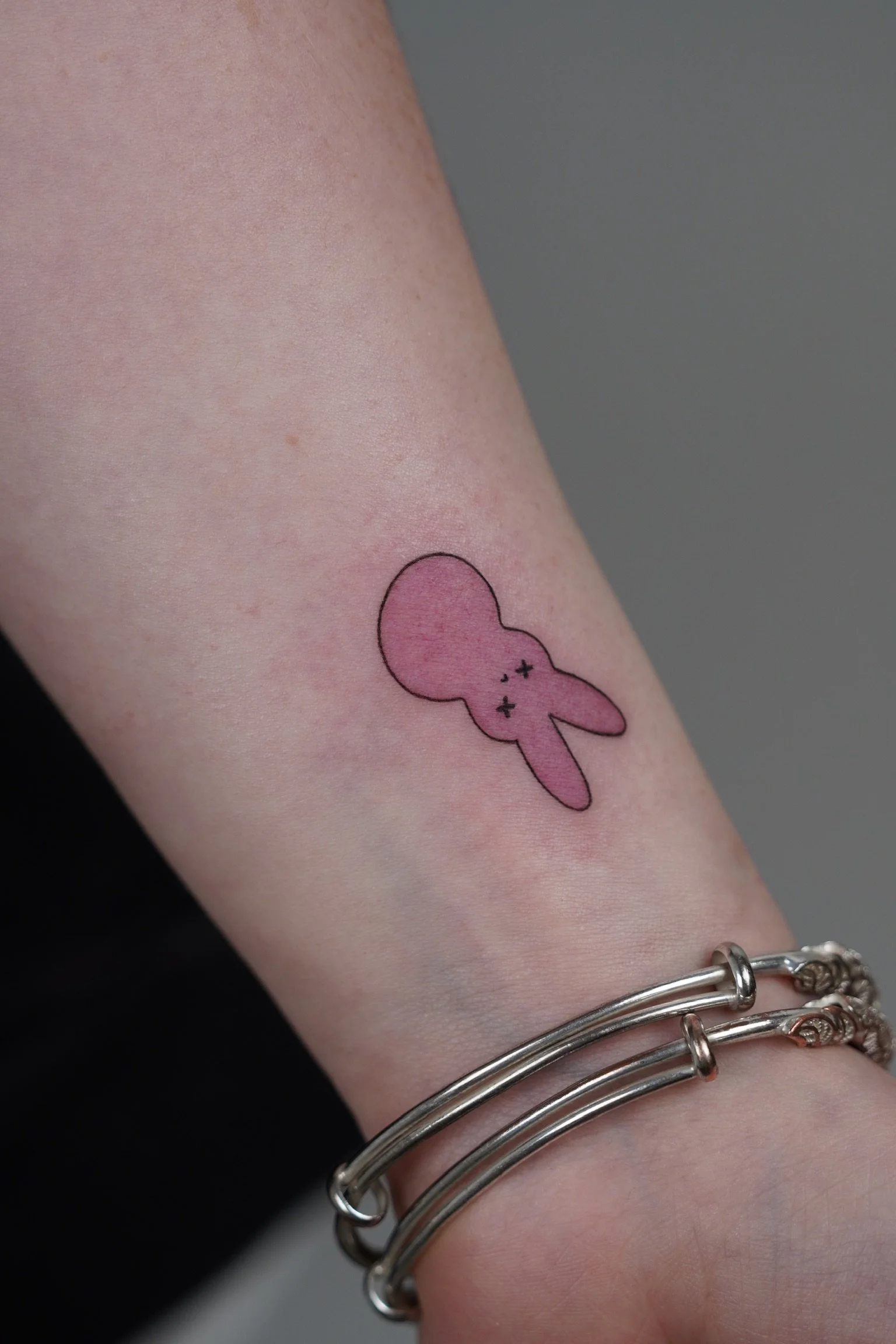 A pink tattoo of a cartoon bunny with crossed-out eyes on a person's forearm, with two silver bracelets.