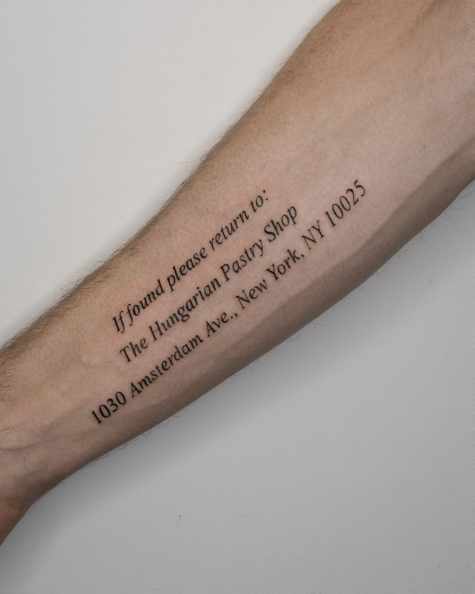 Tattoo on a forearm with an address and instructions for returning a lost item, reading: "If found please return to: The Hungarian Pastry Shop 1030 Amsterdam Ave., New York, NY 10025"