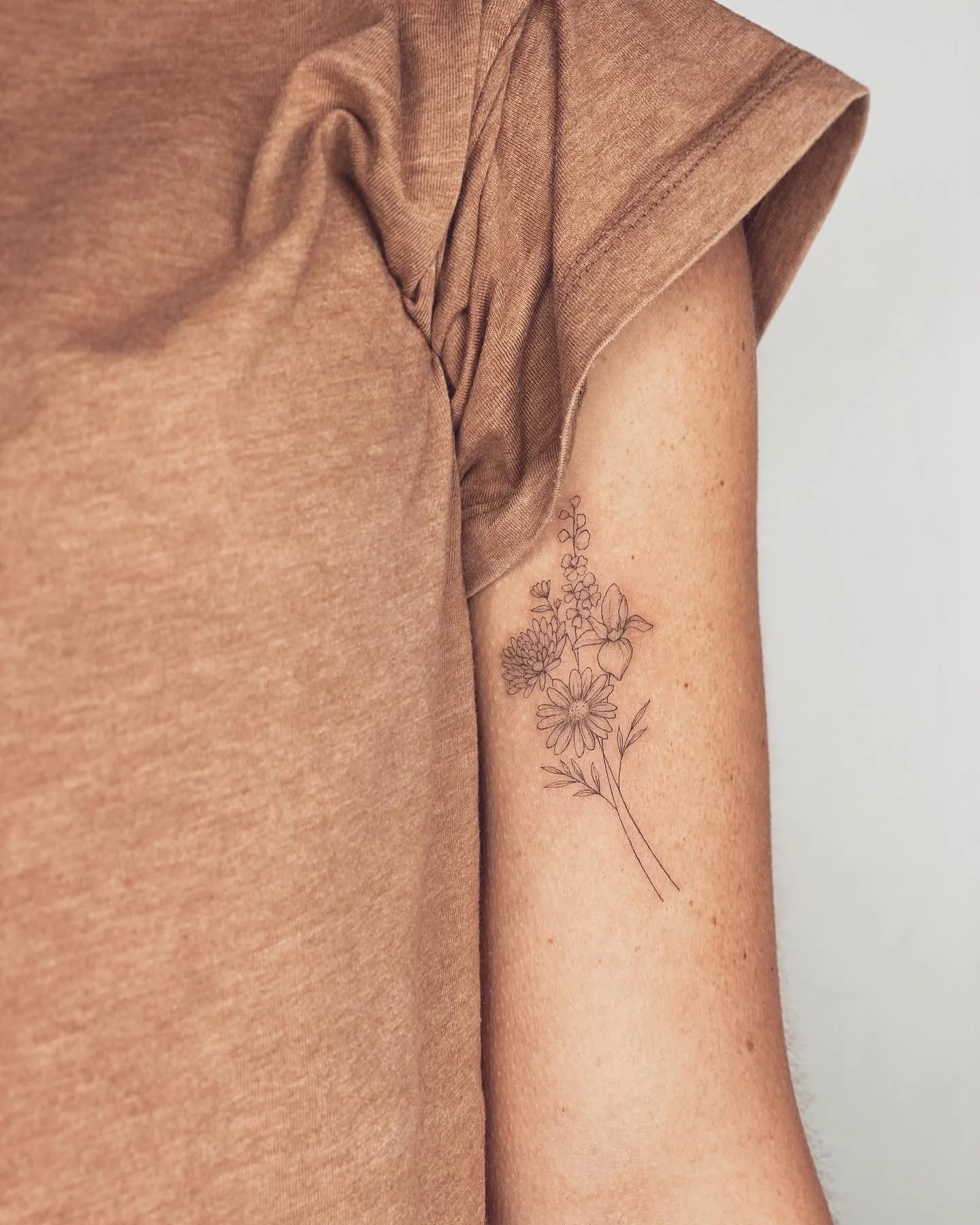 A close-up of a person's upper arm showing a delicate black line art floral tattoo on the inner part of the arm, near the armpit, wearing a brown short-sleeved shirt.