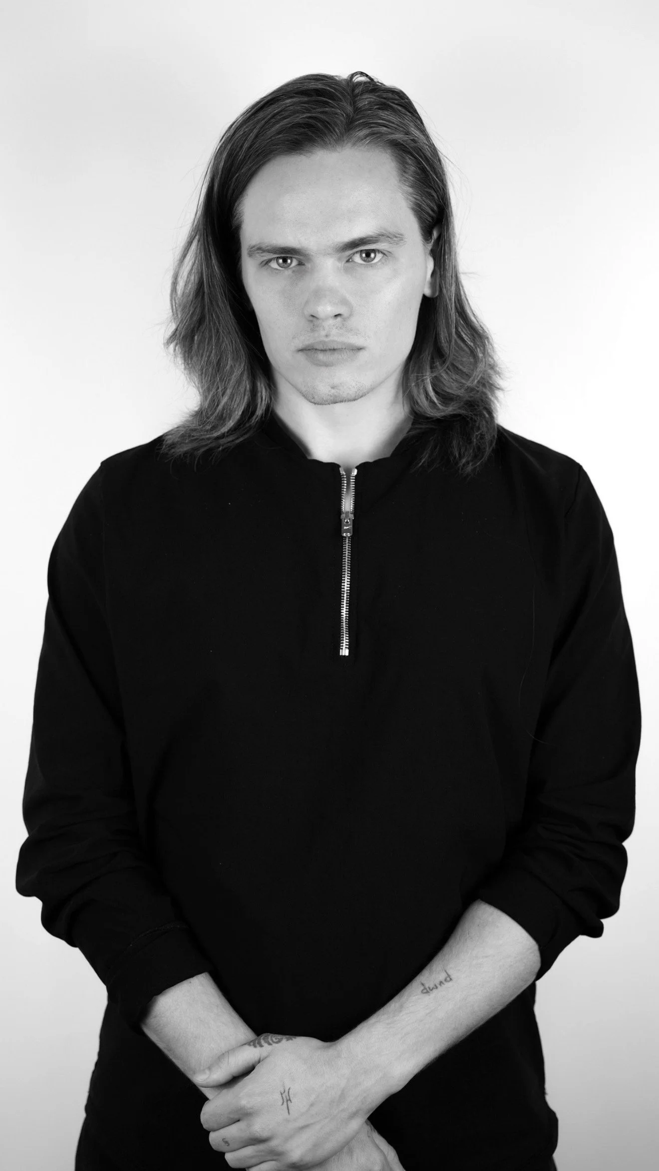 Black and white photo of a young man with long hair, wearing a dark zip-up top, looking directly at the camera with a serious expression.
