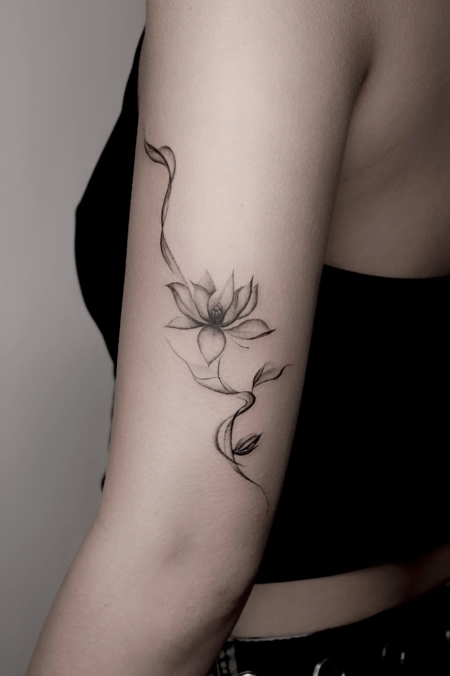 Close-up of a person's upper arm with a black and gray watercolor-style tattoo of a lotus flower and foliage.