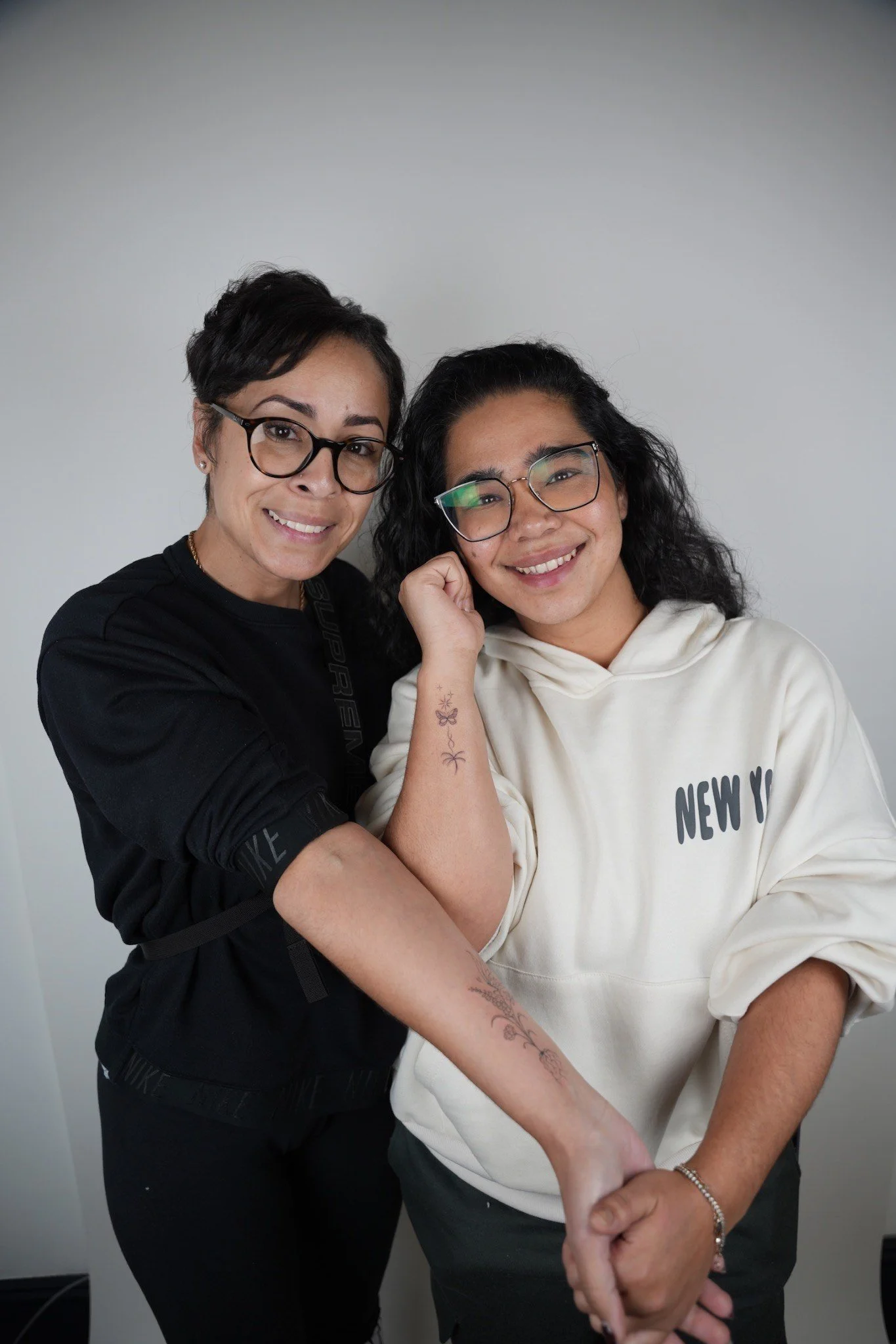 Two women smiling and posing together against a plain wall. One woman has short dark hair, wears glasses, and is dressed in black. The other has long curly hair, glasses, and is dressed in a cream-colored hoodie that says 'New York'.
