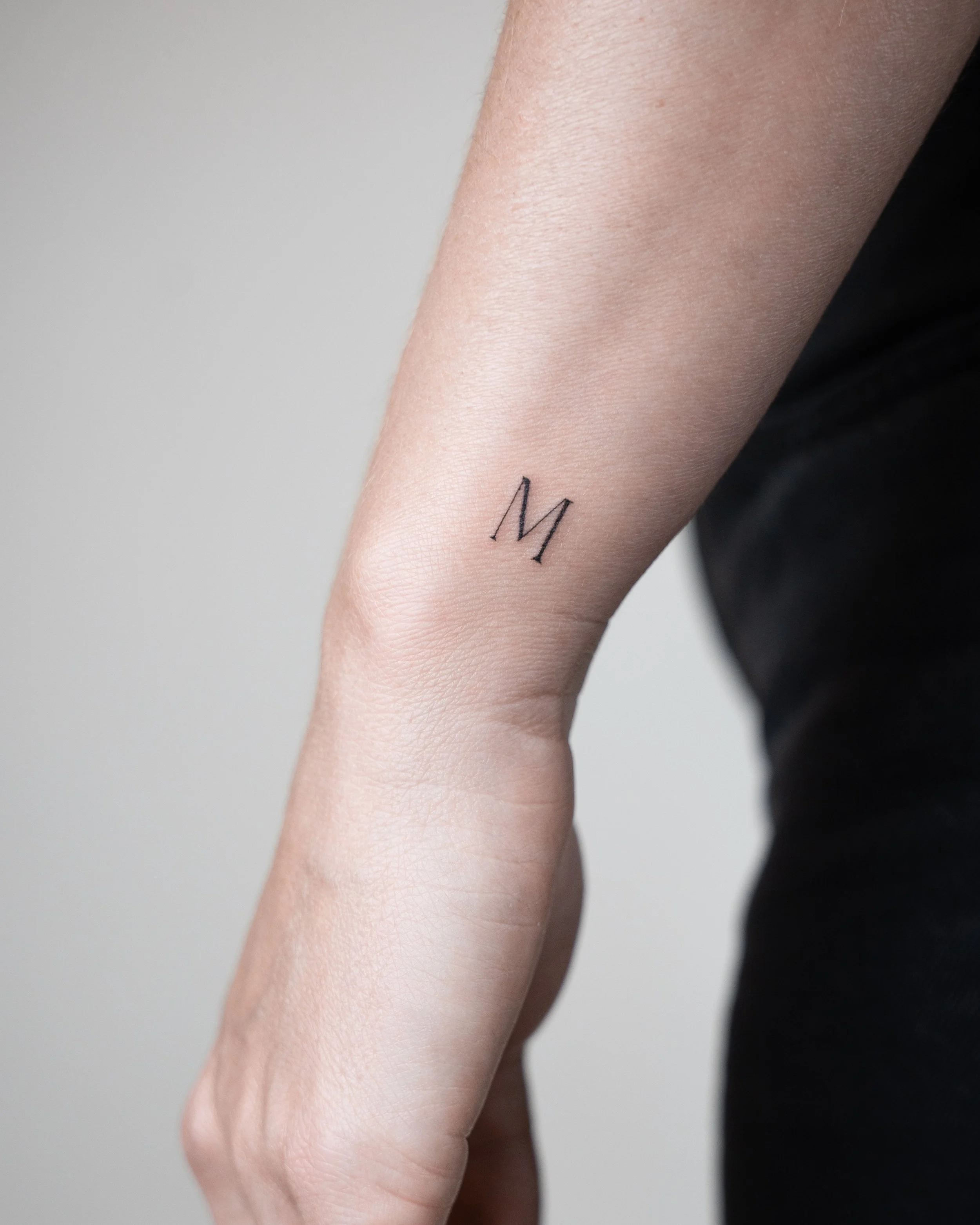 Close-up of a person's inner wrist with a small black tattoo of the letter 'M'.
