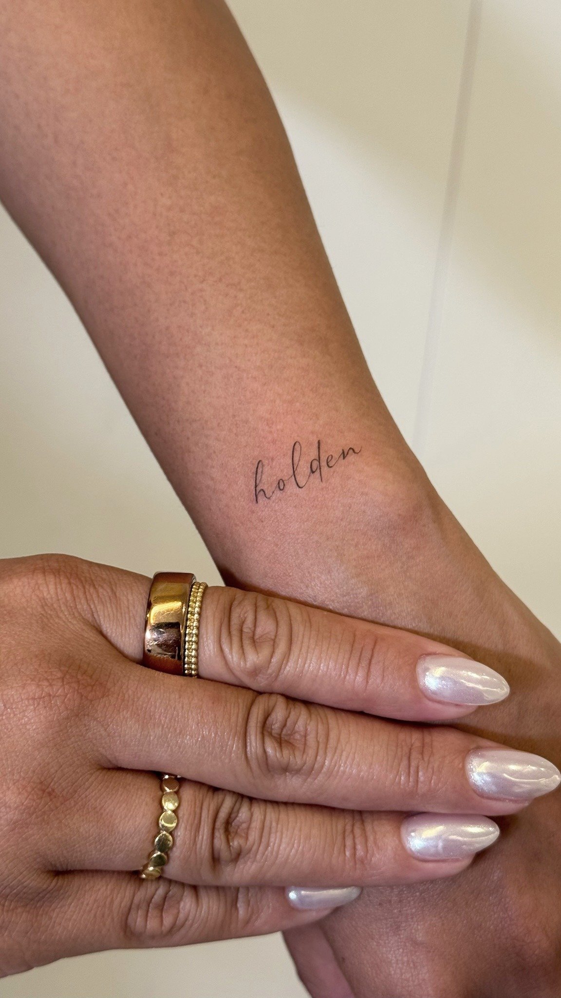 A person's arm with a small tattoo that says 'holden' in cursive. The person is wearing multiple rings and has painted nails.