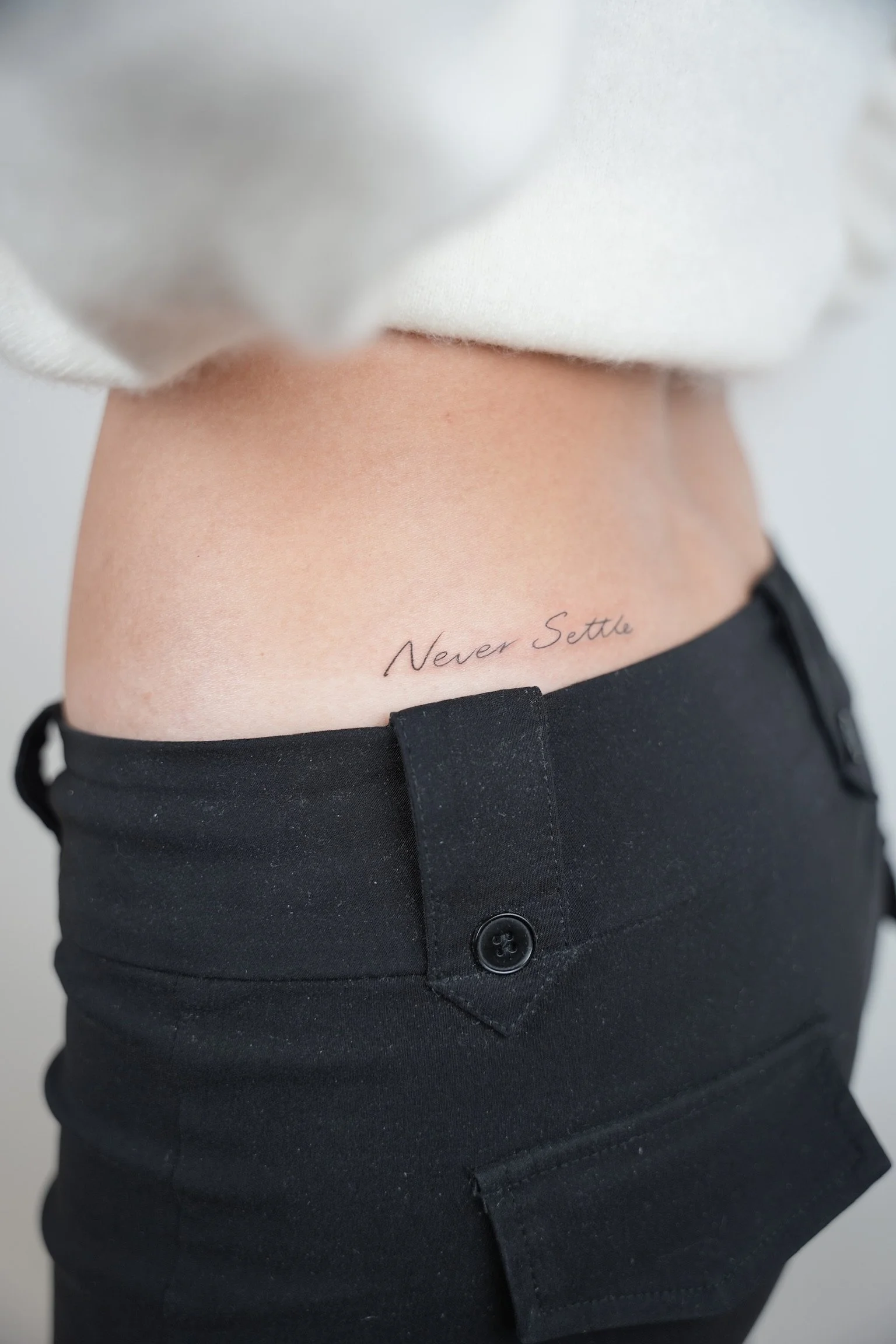 Close-up image of a person's midsection showing a tattoo that says "Never Settle" on the lower abdomen, with the person wearing black pants and a white top.