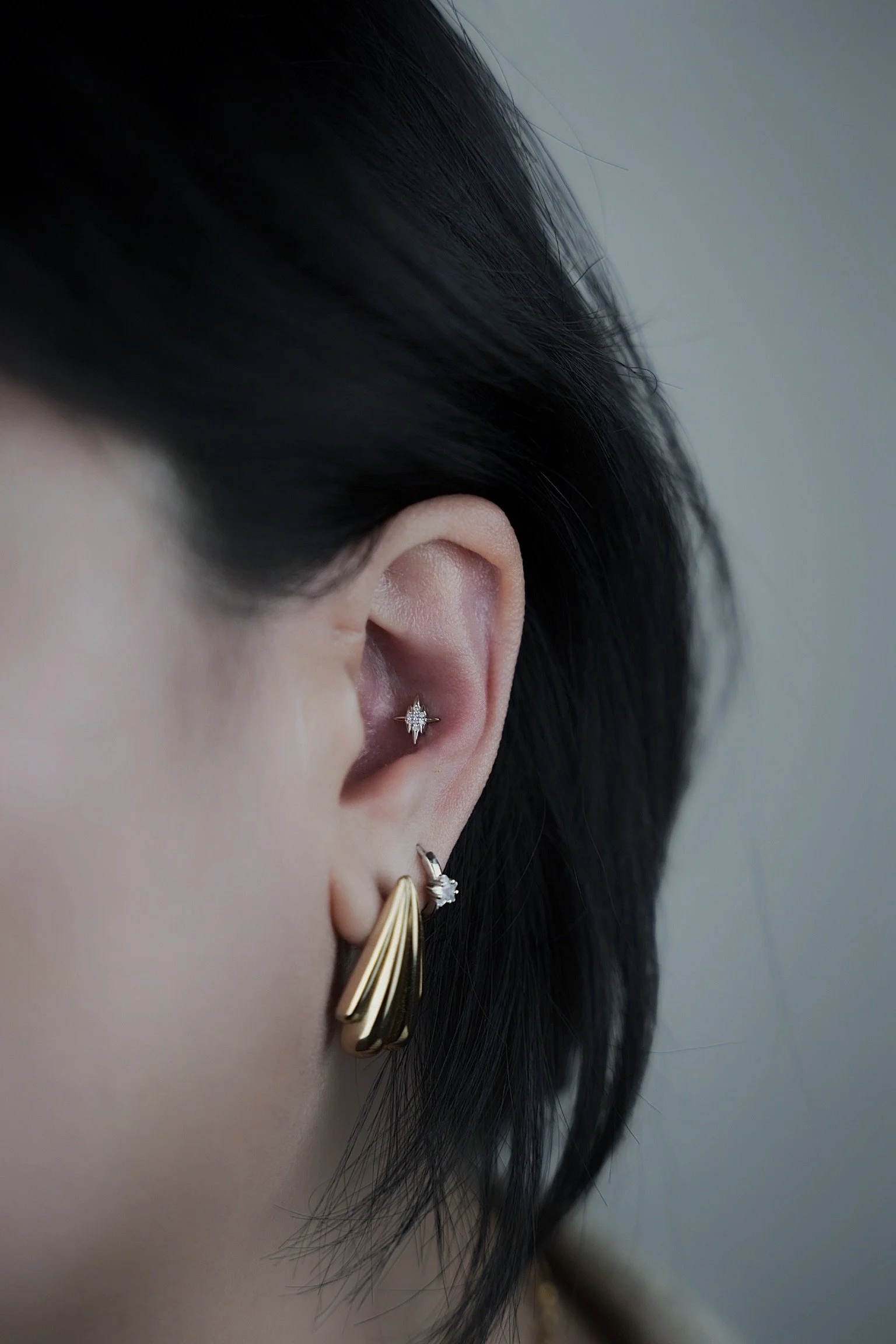 Close-up of a person's ear with multiple earrings, including a gold hoop, a silver star-shaped stud, and a small diamond-shaped stud with a black gemstone, with dark hair partially covering the ear.
