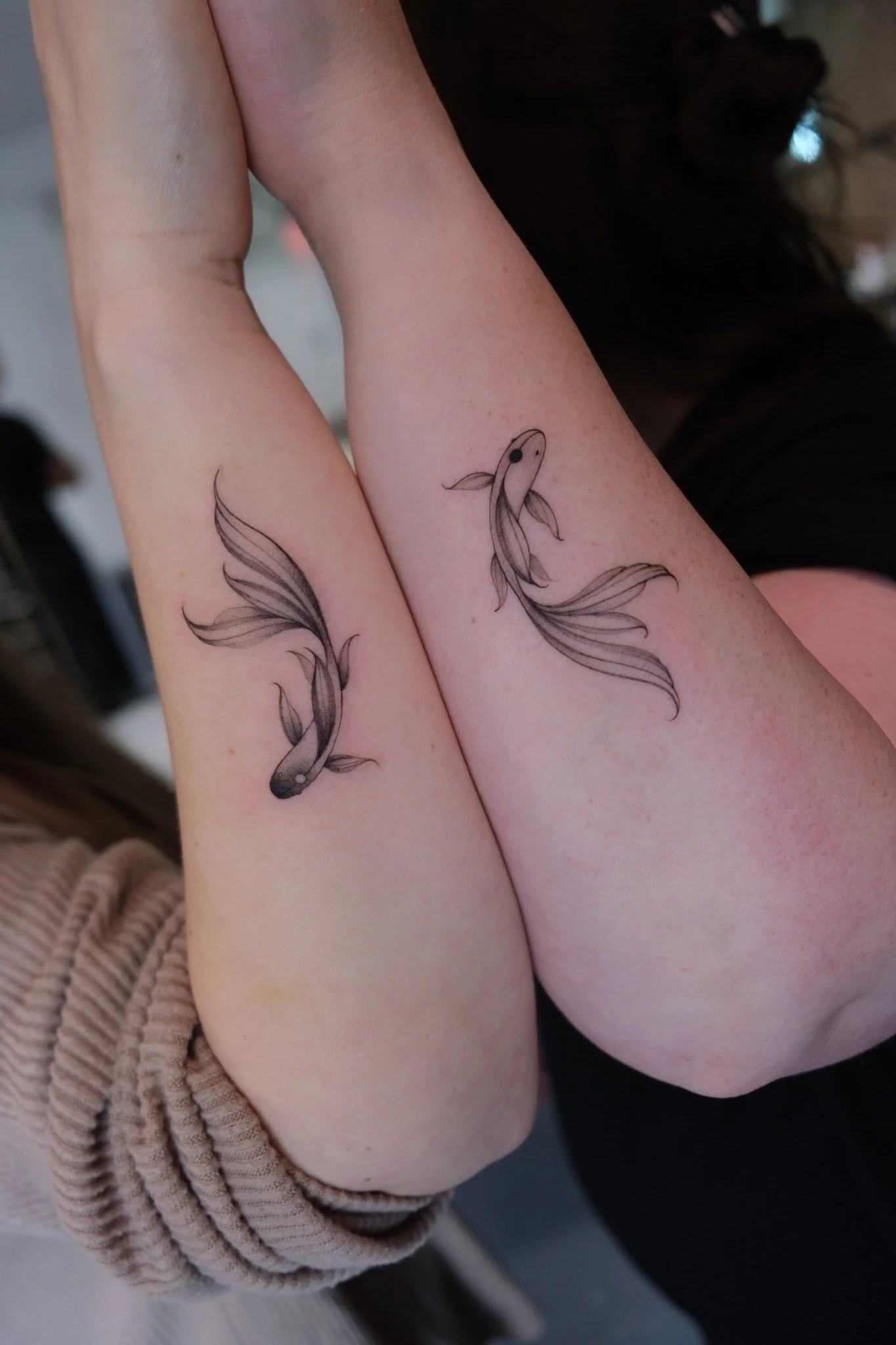 Close-up of two forearms with matching black line art koi fish tattoos with flowing fins and tails.