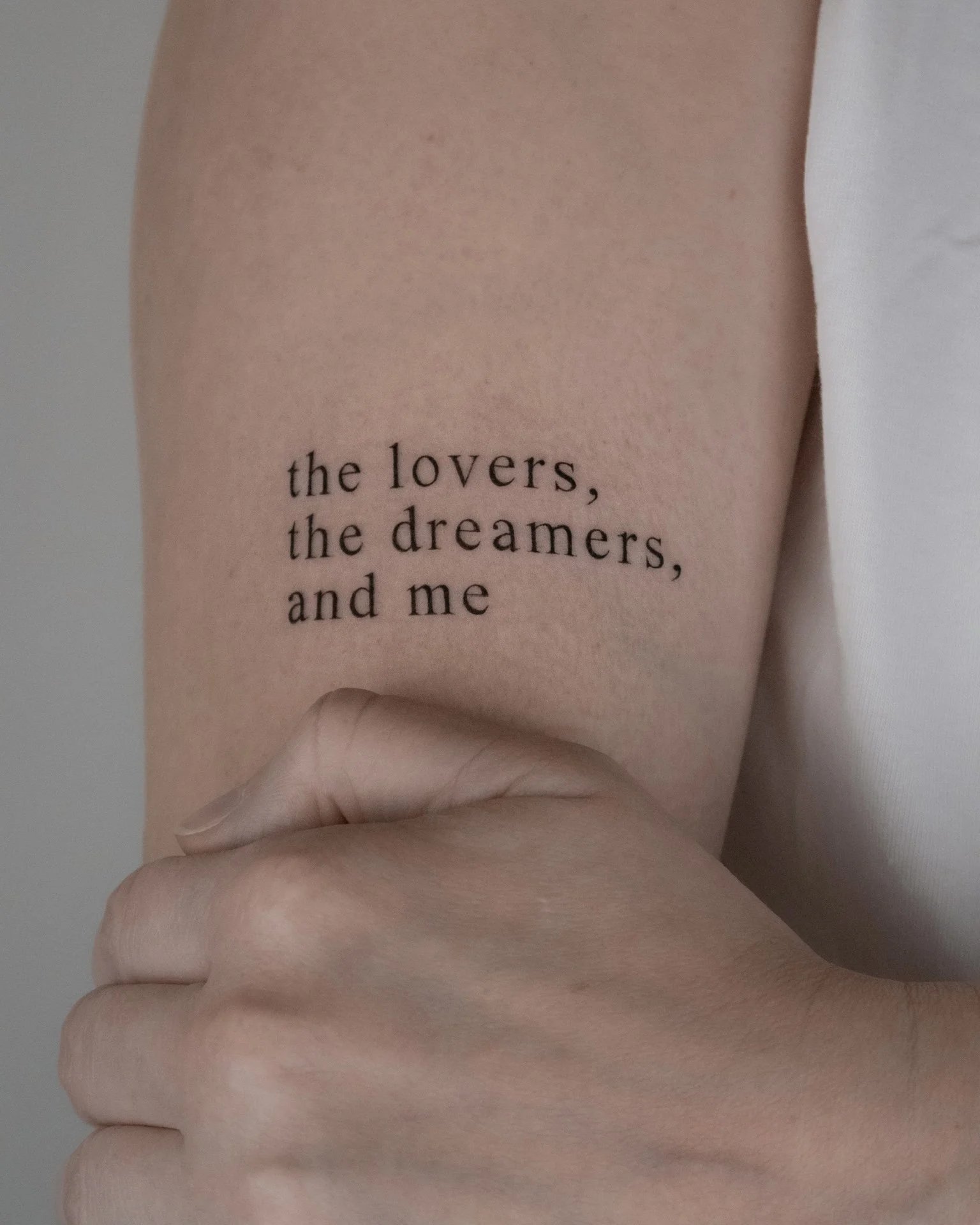 A close-up of a person's arm with a tattoo that reads, 'the lovers, the dreamers, and me' in black text.
