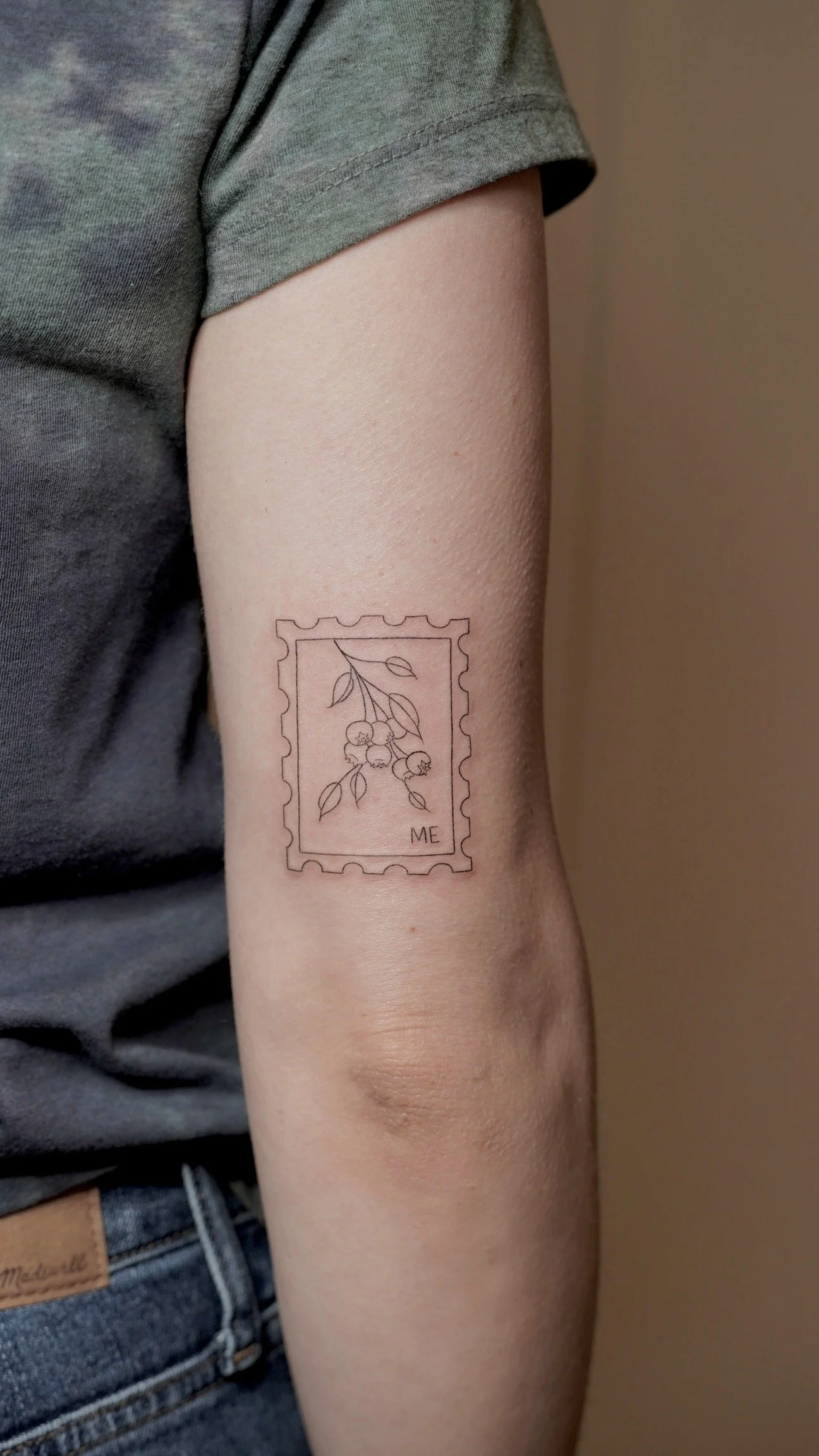 A tattoo of a postage stamp with a line drawing of a branch with small round berries and leaves, with the word "ME" in the bottom right corner, on a person's upper arm.