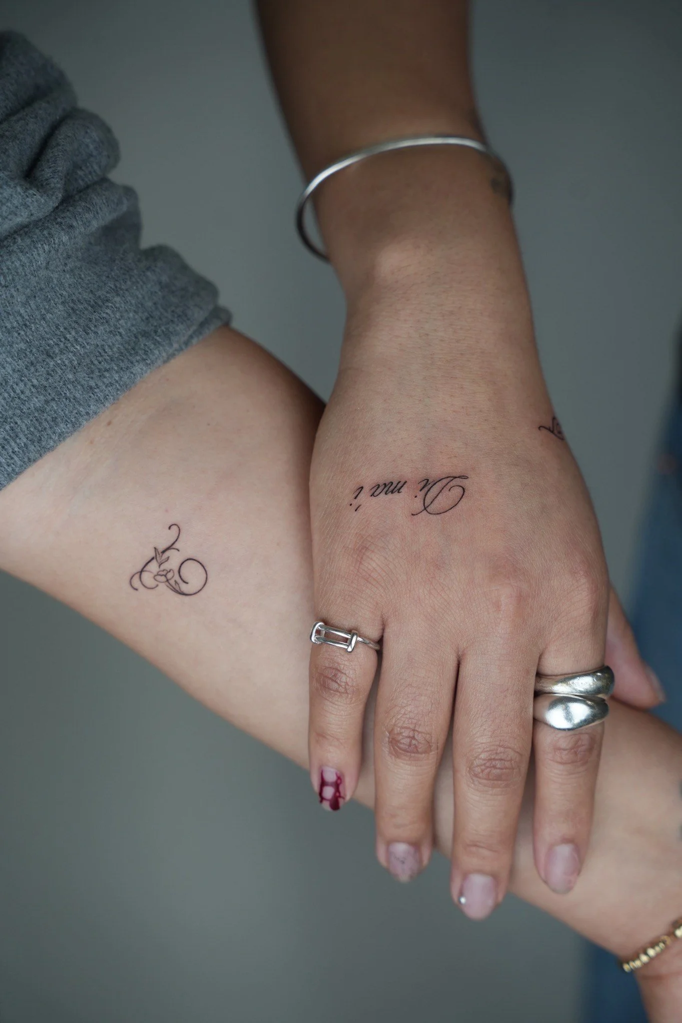 Close-up photo of two hands gently resting on top of each other, displaying tattoos with cursive writing, various rings, and a simple bracelet on a gray background.