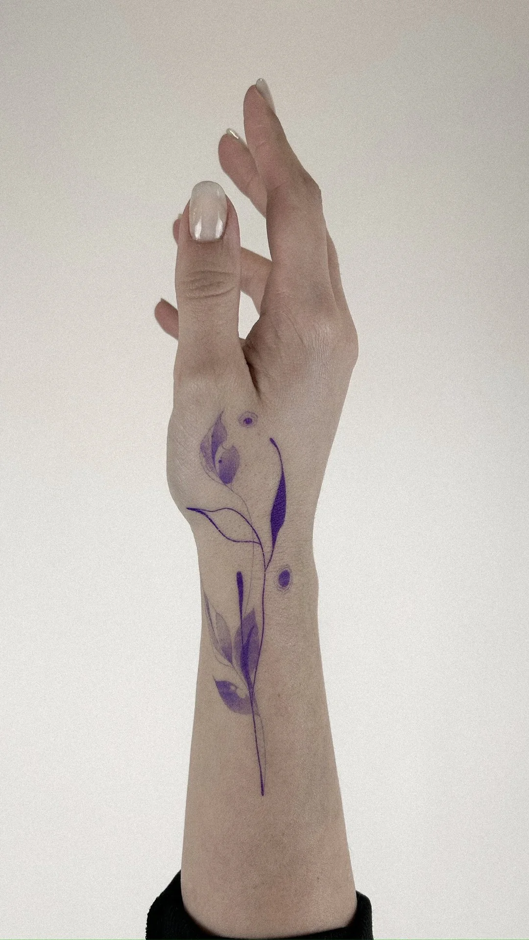 A person's hand with a purple and black floral tattoo on the wrist and forearm, with manicured nails painted in a pearly white color.