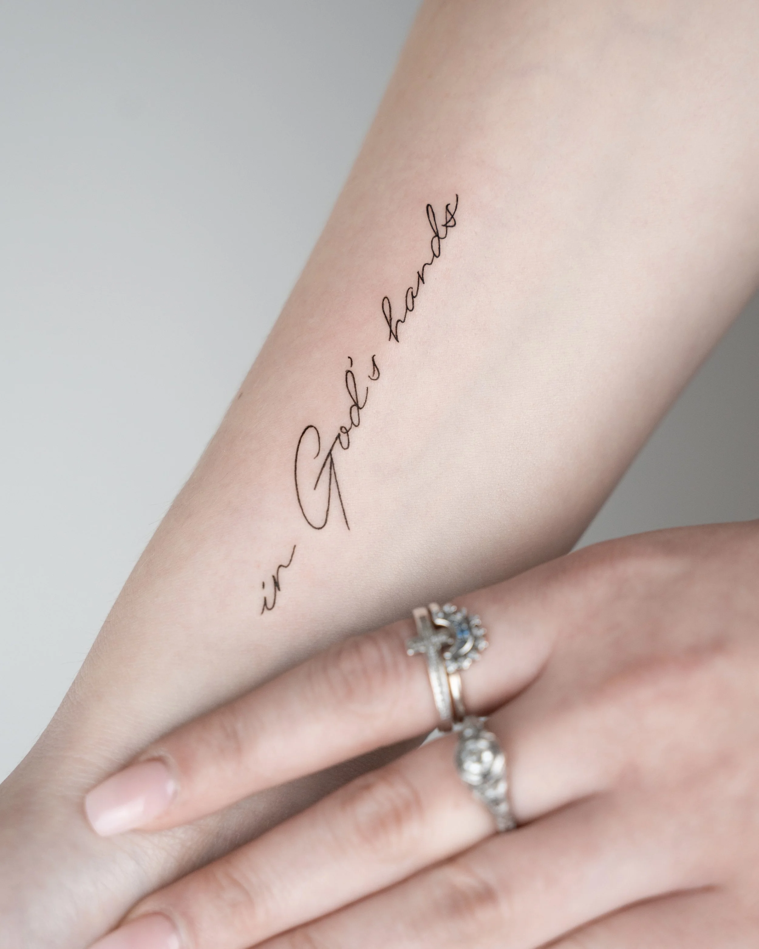 A close-up of a person's forearm with a tattoo that reads 'in God's hands' in cursive script. The person is wearing two rings on their finger, one with a cross design and the other with a small stone. The background is plain and light-colored.