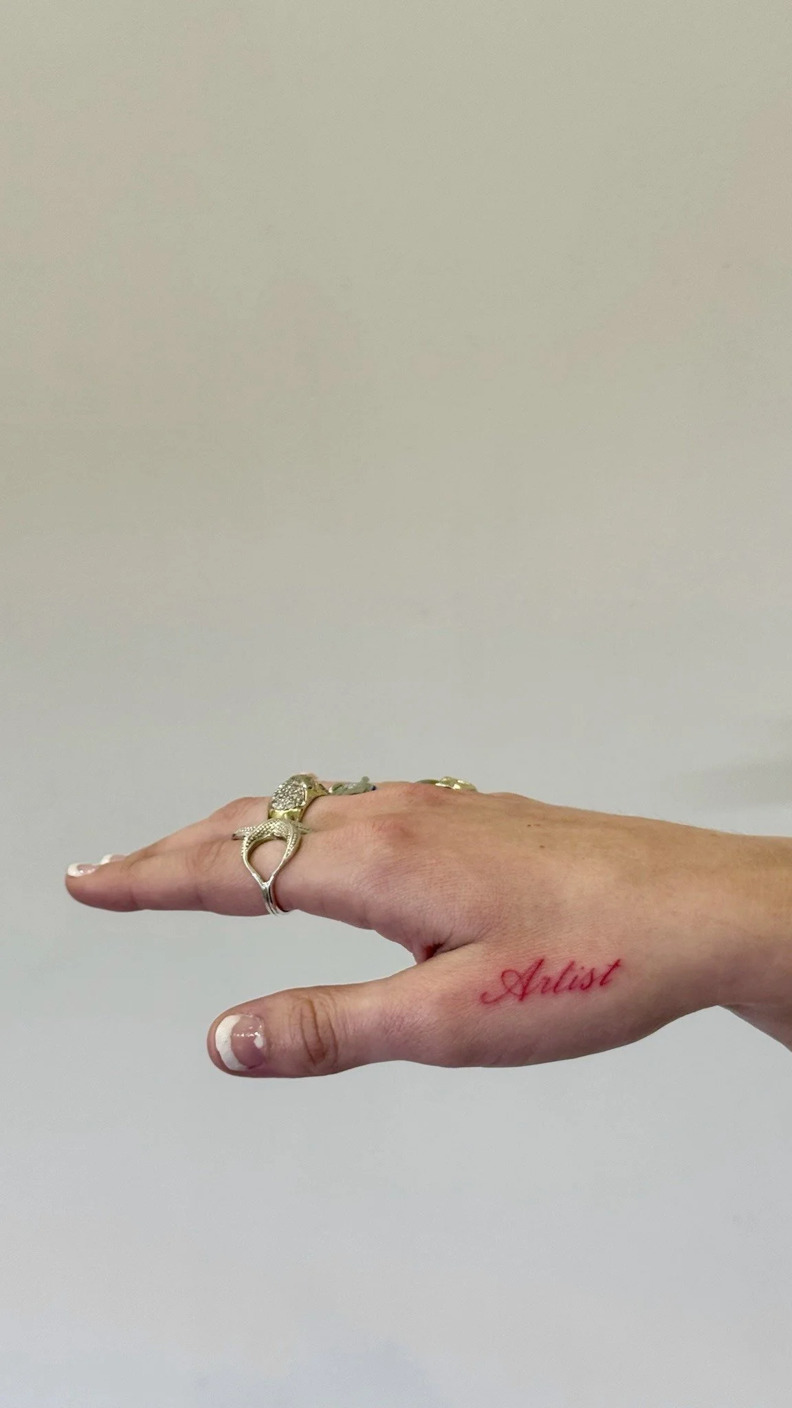 A hand with rings and a red tattoo that says 'Artist' against a plain wall.