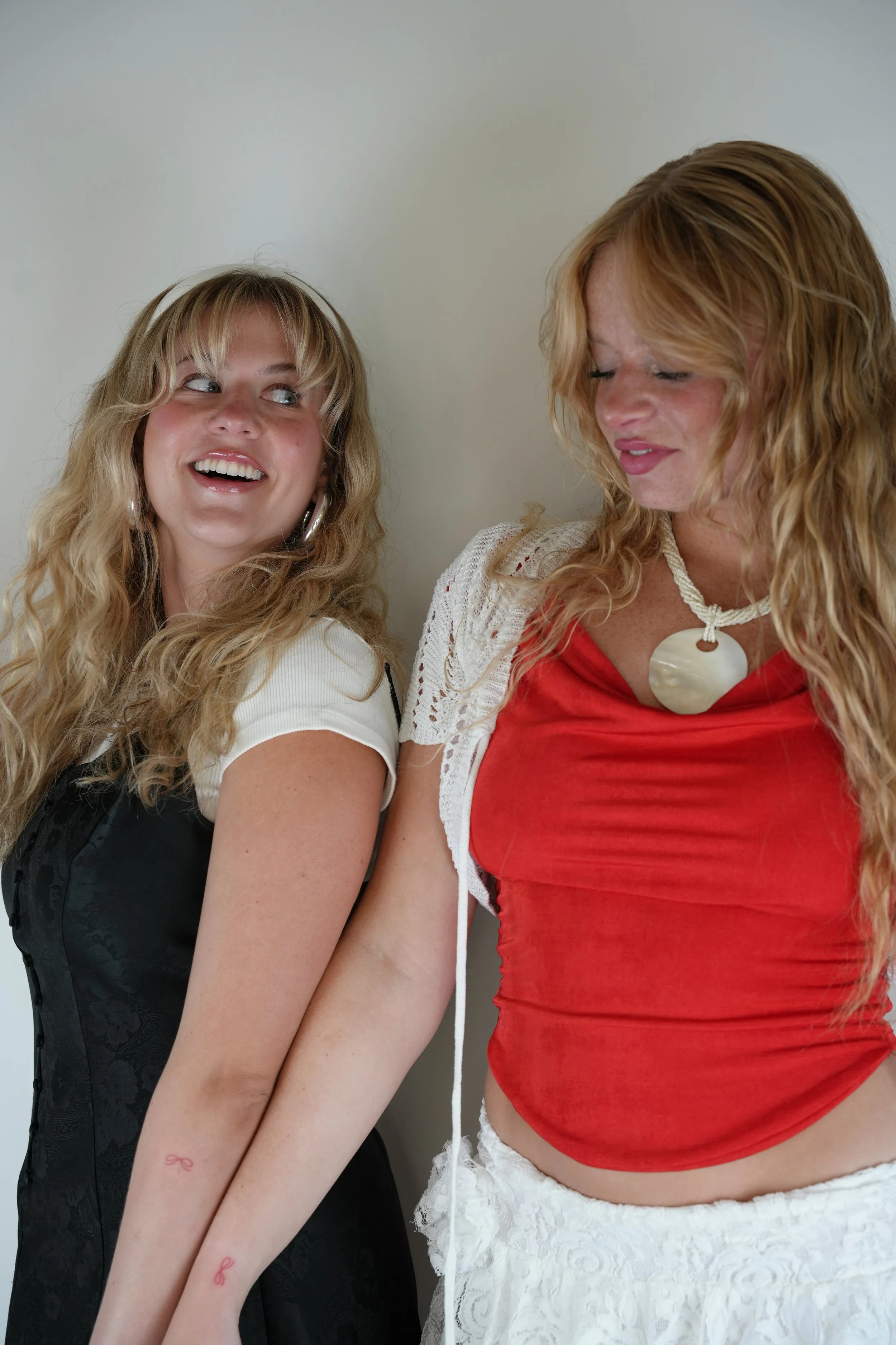 Two women with long, curly blonde hair standing close together, smiling and looking at each other against a plain light-colored wall.
